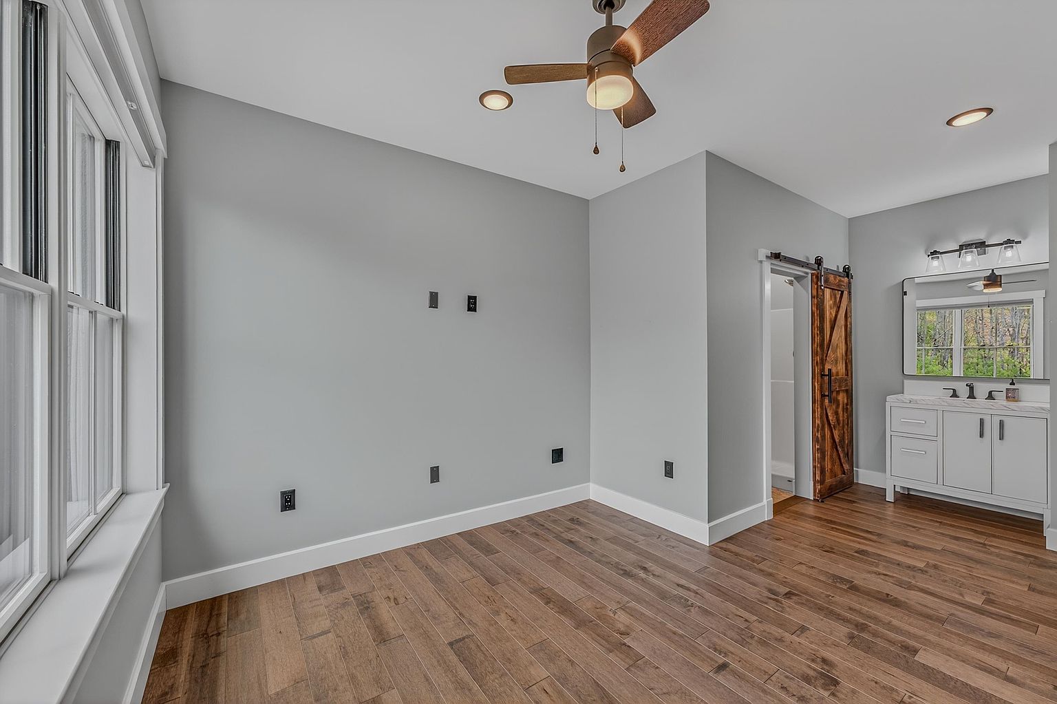 This primary bedroom features light gray walls, warm-toned wood flooring, and a ceiling fan with integrated lighting. A sliding barn door provides access to an adjoining bathroom, which includes a white vanity and a mirror. The room is brightened by large windows on the left, creating an open and airy atmosphere.