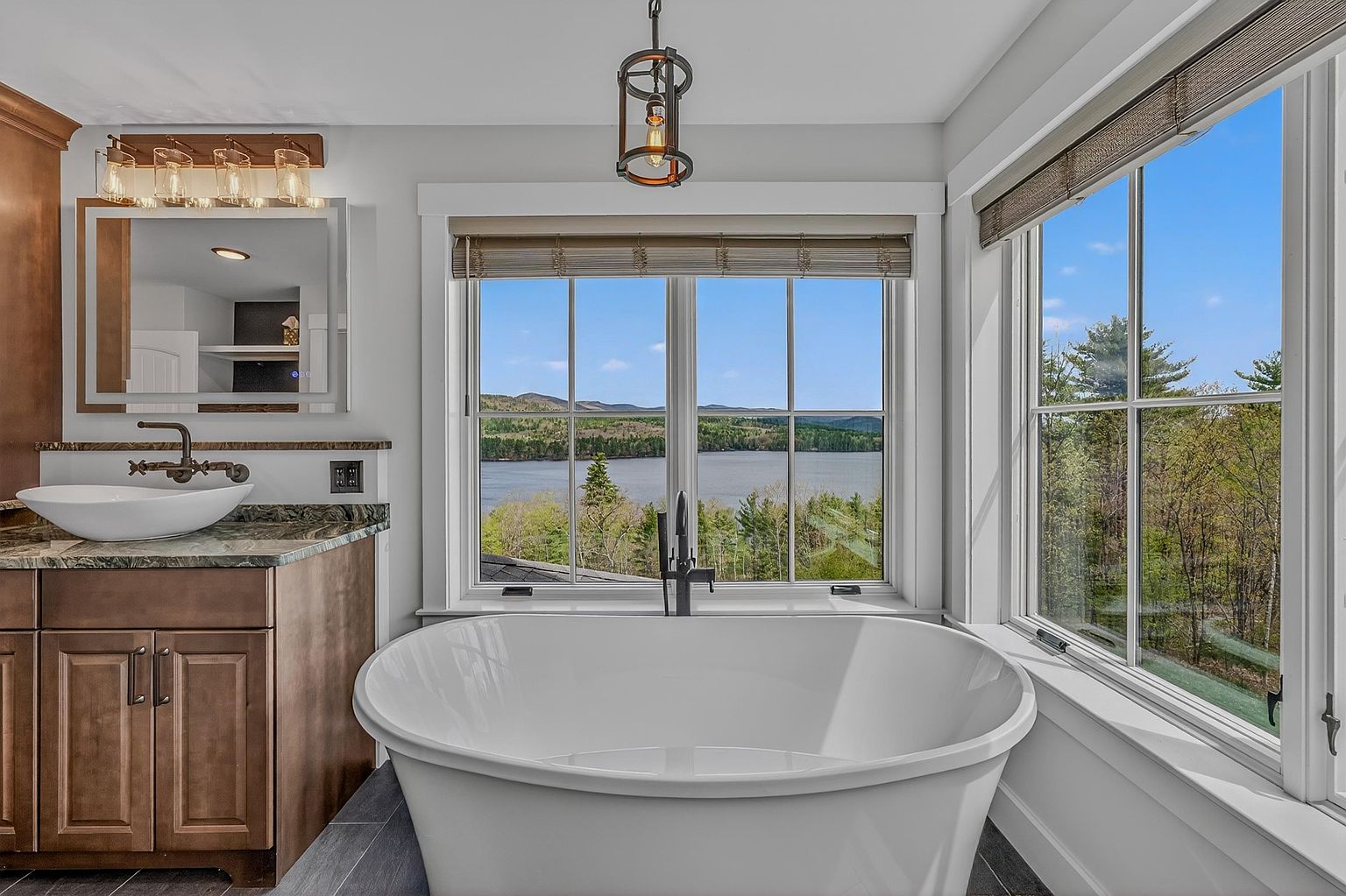 This primary bathroom features a deep, freestanding soaking tub positioned directly in front of large windows that offer a panoramic view of a lake and surrounding forest. To the left, a vanity with a vessel sink and stone countertop is complemented by a modern mirror and warm lighting fixtures. The space combines a clean, contemporary aesthetic with natural elements, creating a serene and spa-like atmosphere.