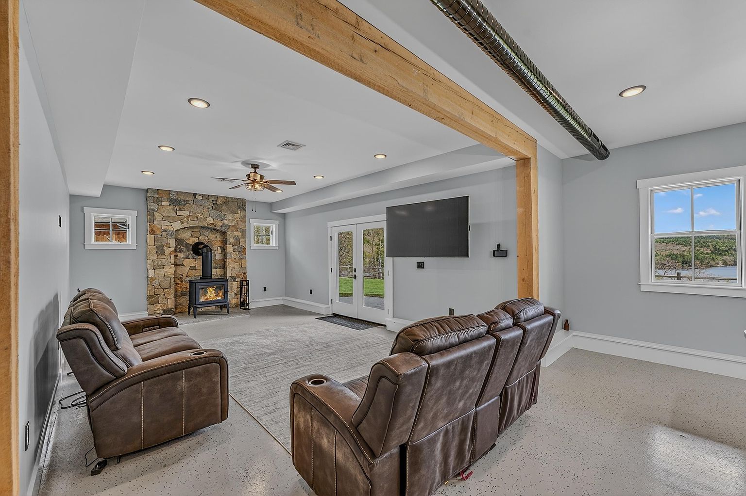 This spacious living room features a cozy wood-burning stove set against a prominent stone-clad chimney breast, creating a rustic focal point. The room is furnished with two brown leather sofas arranged on a large area rug, while a wall-mounted television provides entertainment. A large wooden beam spans the ceiling, and double glass doors lead to the outdoors, allowing natural light to fill the space.