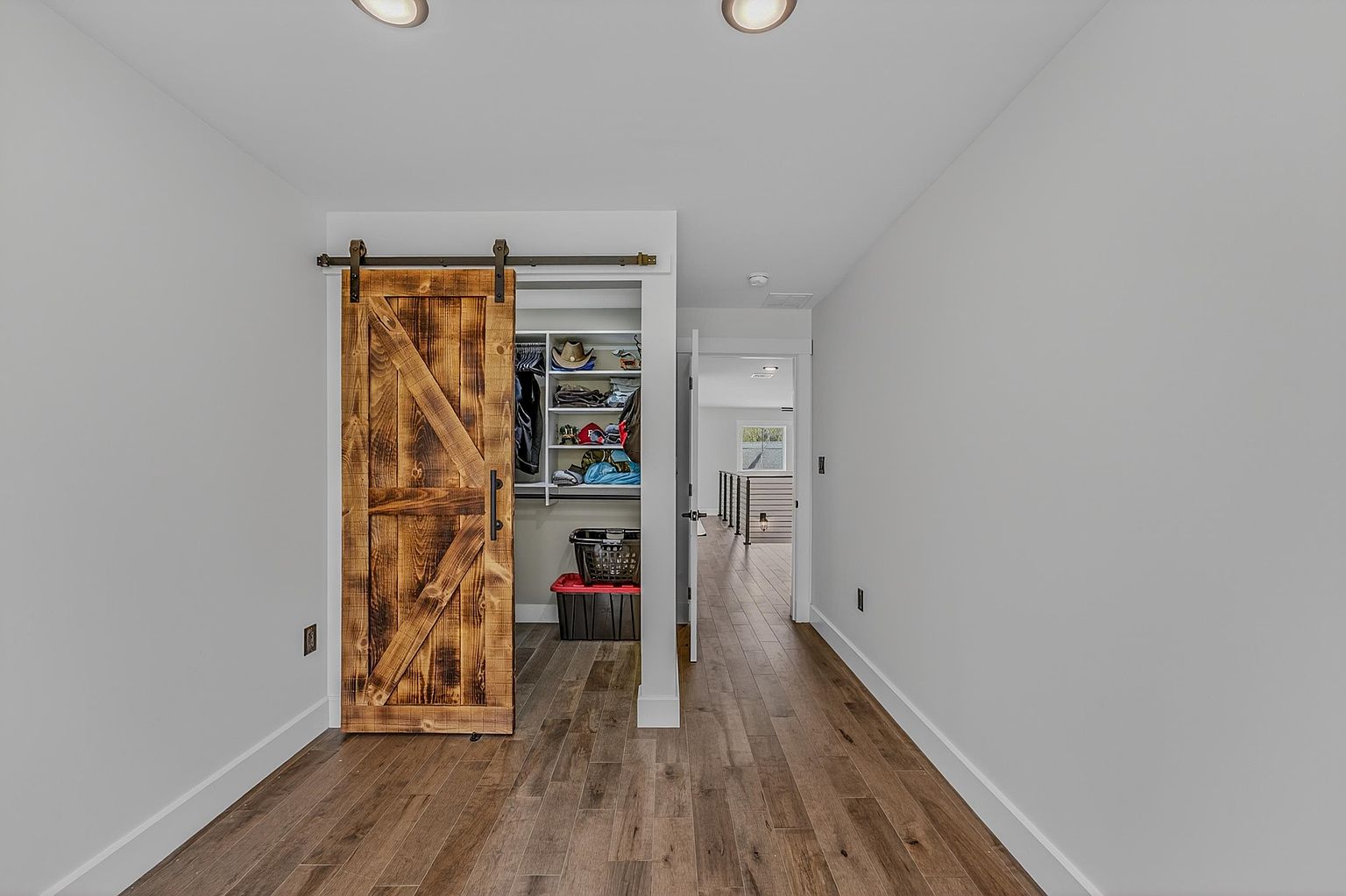 This interior view features a walk-in closet accessed by a rustic, sliding barn-style door with a dark metal track. The closet interior is organized with white shelving and hanging rods, while the surrounding room showcases light-toned walls and wood-look flooring. A doorway in the background leads to an adjacent hallway, providing a sense of depth and flow within the home's layout.