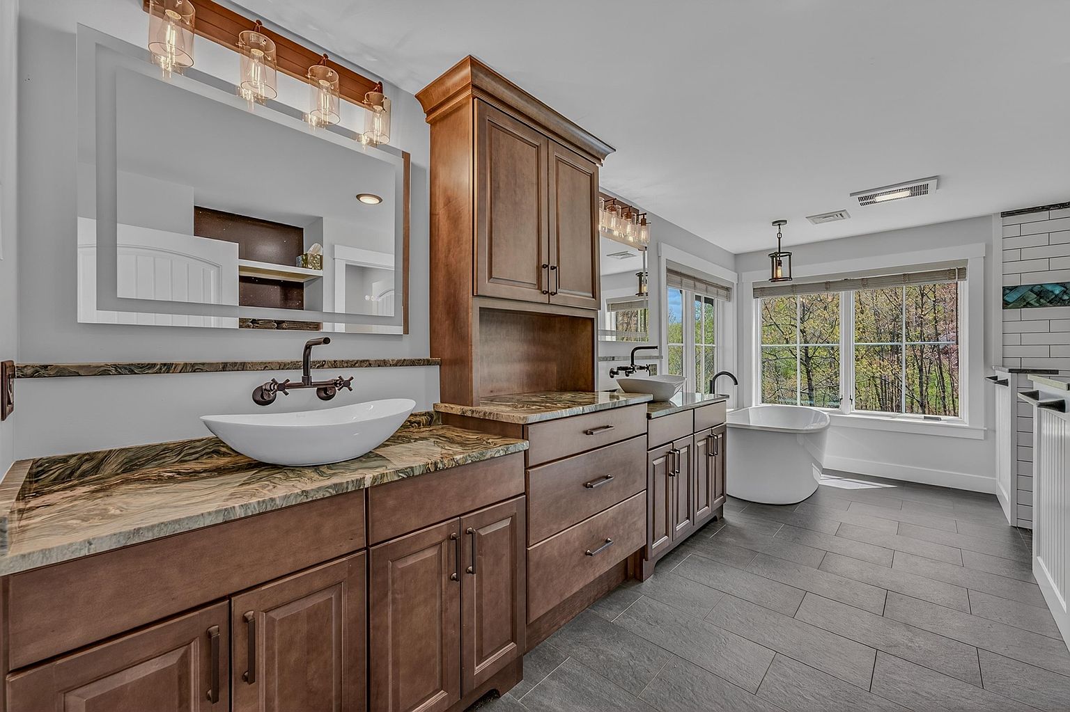 This spacious primary bathroom features a dual-vanity setup with dark wood cabinetry and patterned stone countertops. Two vessel sinks are paired with wall-mounted faucets, while a freestanding soaking tub sits near large windows that provide ample natural light and a view of the outdoors. The room is finished with dark tile flooring and modern lighting fixtures, creating a sophisticated and serene atmosphere.
