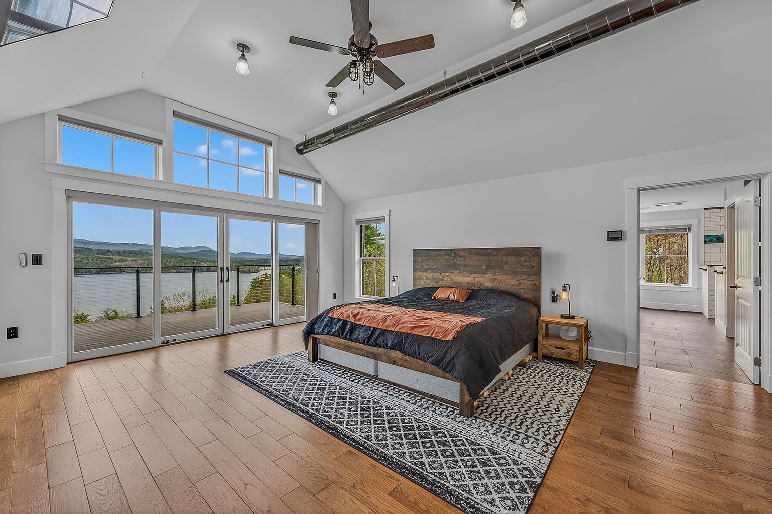 This spacious primary bedroom features a vaulted ceiling with an exposed industrial-style duct and a central ceiling fan. A large bed with a rustic wood headboard sits atop a patterned area rug, positioned to face a wall of expansive glass sliding doors that provide panoramic views of the lake and mountains. The room is finished with light wood flooring and leads into an adjacent hallway, creating an open and airy atmosphere.