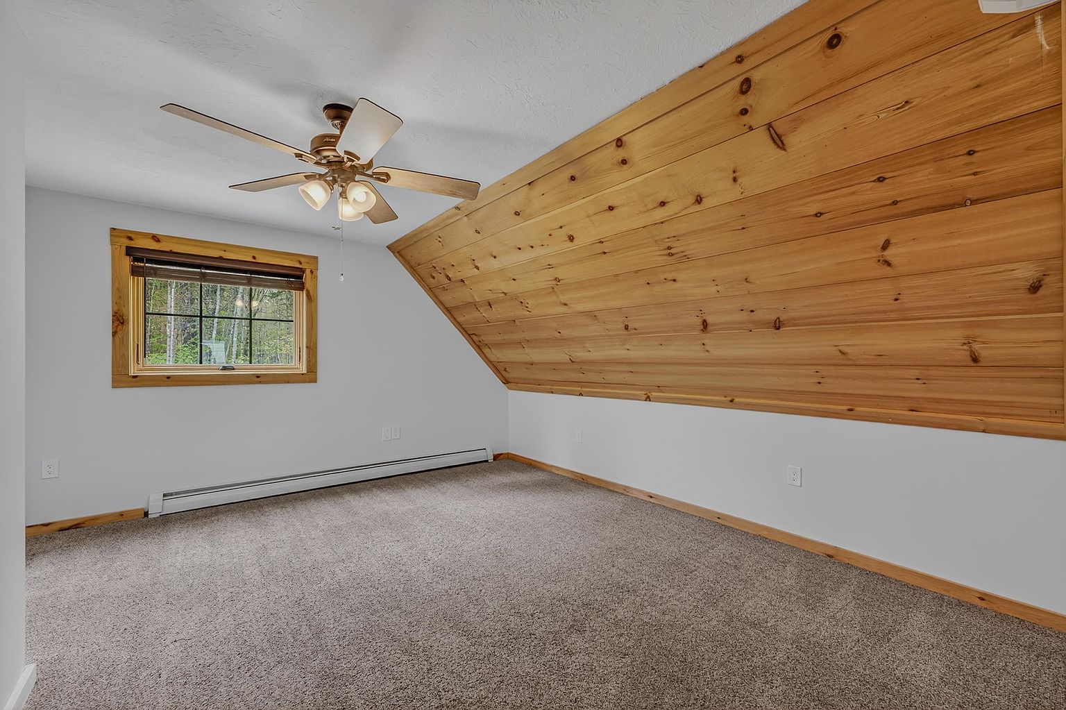 This attic bedroom features a distinctive sloped ceiling paneled in light wood, creating a cozy, rustic atmosphere. The room is carpeted in a neutral tone and includes a window with a wood-trimmed frame, a ceiling fan with integrated lighting, and baseboard heating. The space offers a clean, blank canvas with white walls, ideal for a guest room or a quiet retreat.