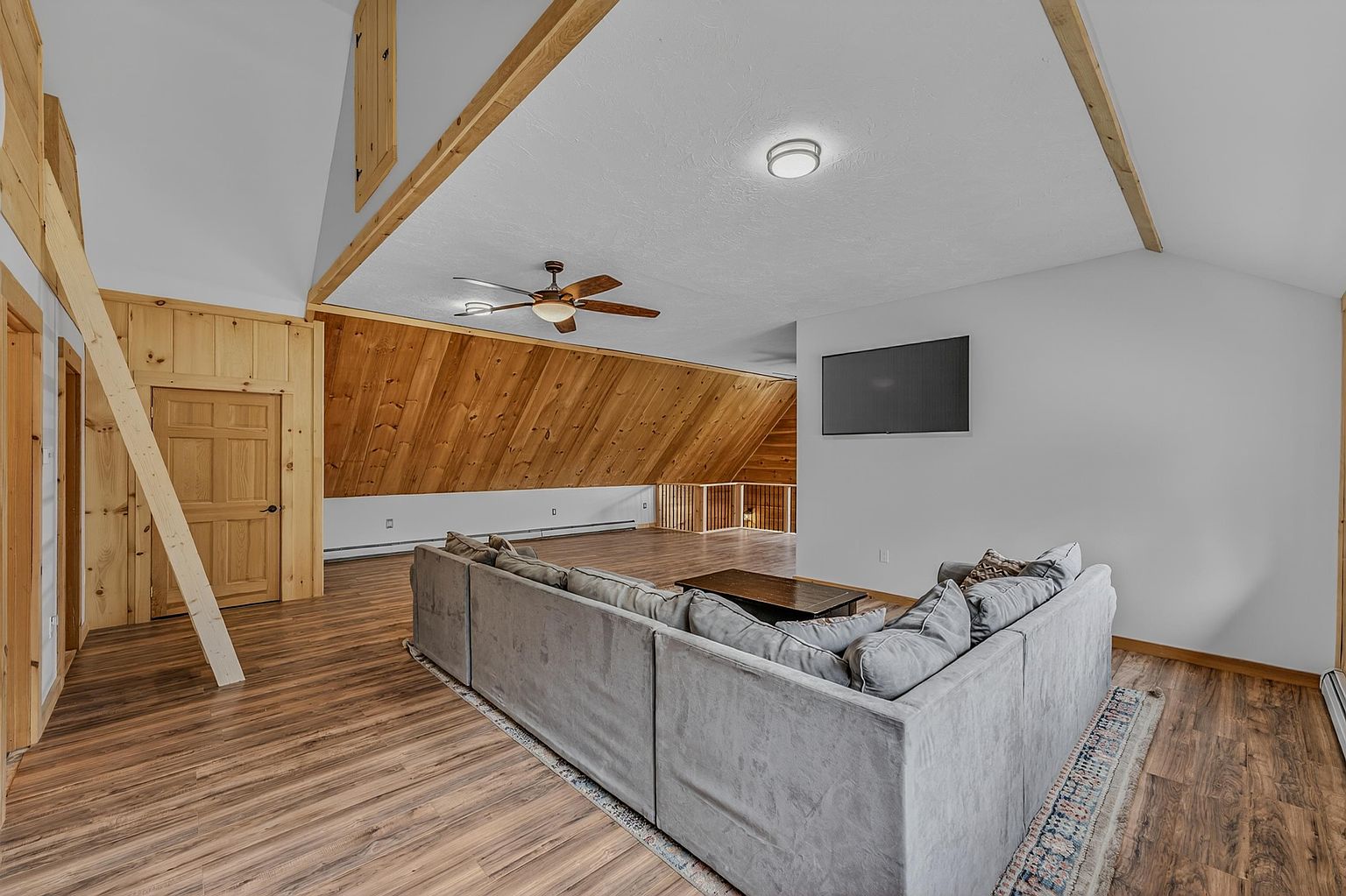 This spacious living area features a vaulted ceiling with exposed wooden beams and a large, light-colored sectional sofa positioned to face a wall-mounted television. The room showcases a rustic aesthetic with wood-paneled walls and warm-toned wood-look flooring, creating an open and inviting atmosphere. A ceiling fan provides air circulation, while the open layout flows into a secondary space with a wooden railing overlooking the lower level.