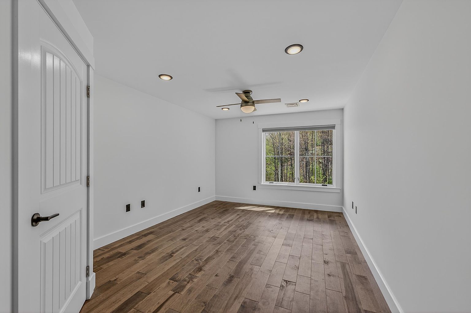 This bright, neutral-toned bedroom features warm-toned wood-look flooring and crisp white walls, creating a clean and versatile living space. A ceiling fan with integrated lighting is centered in the room, while recessed lighting provides additional illumination. A large window on the far wall allows for natural light and offers a view of the surrounding trees.