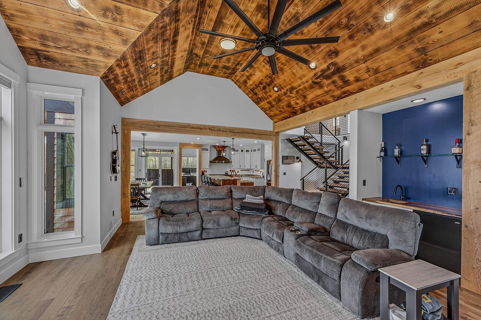 This spacious open-concept living area features a vaulted ceiling with rustic wood planking and a large, modern ceiling fan. A plush, grey sectional sofa sits atop a textured area rug, facing toward the open kitchen and dining area in the background. To the right, a built-in wet bar with a deep blue accent wall and glass shelving adds a sophisticated touch, while a staircase with metal railings leads to the upper level.