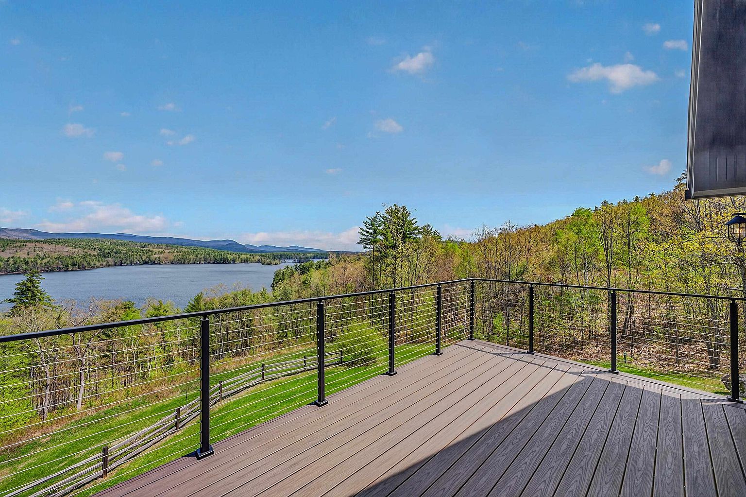 This elevated deck features modern composite decking and a sleek black metal railing with horizontal cable infill, providing an unobstructed view of the surrounding landscape. The perspective looks out over a lush green lawn, dense forest, and a serene lake nestled against distant mountains under a clear blue sky. The clean lines and open design create an inviting outdoor living space perfect for enjoying the scenic natural environment.