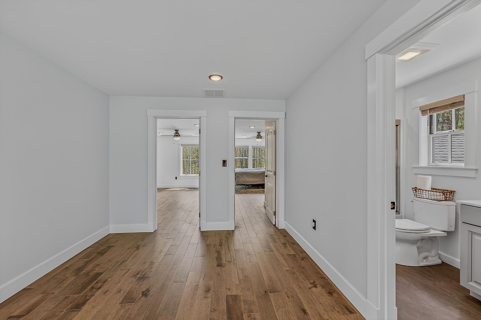 This interior view captures a bright hallway featuring light-toned walls and warm-toned wood flooring. The space provides access to two bedrooms through open doorways and a bathroom on the right, creating a clean and open transition between rooms. The perspective is centered, emphasizing the flow of the home and the consistent design aesthetic throughout the living areas.