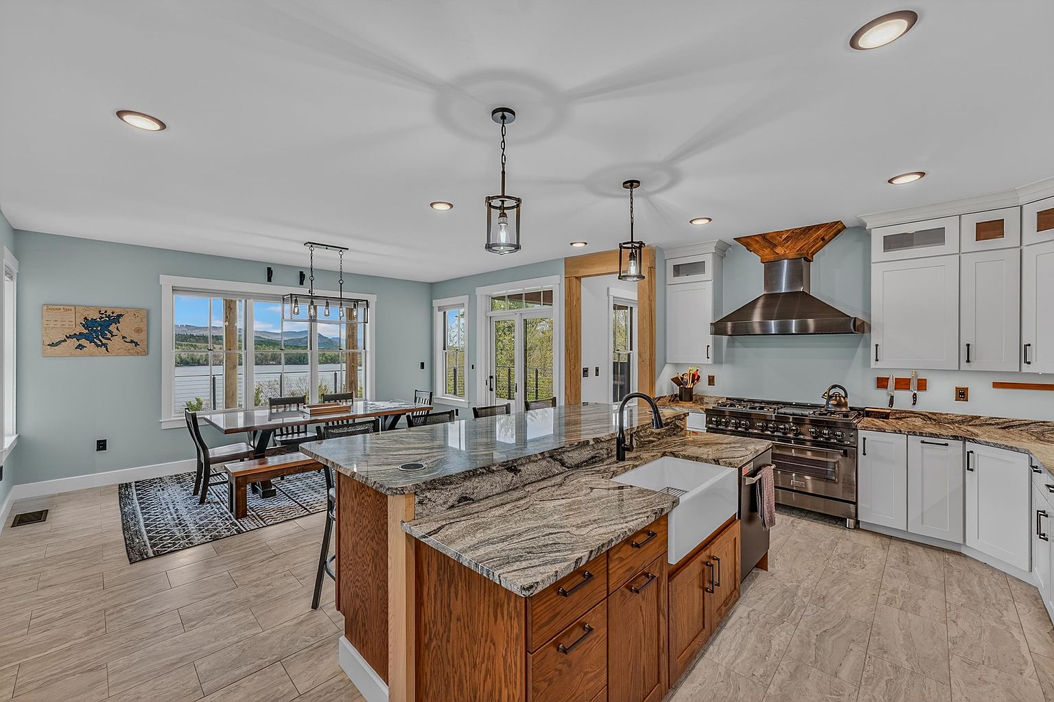 This spacious, open-concept kitchen features a large central island with a farmhouse sink and stone countertops, complemented by white cabinetry and stainless steel appliances. A dining area sits adjacent to the kitchen, framed by large windows that provide a scenic view of the water and mountains beyond. The space is illuminated by a mix of recessed lighting and decorative pendant fixtures, creating a bright and inviting atmosphere.