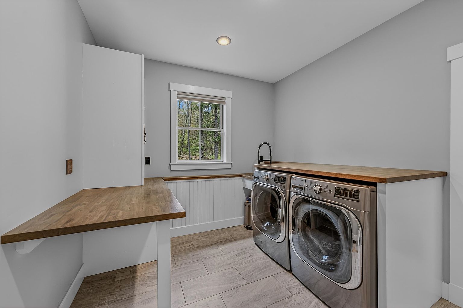 This functional laundry room features a clean, modern aesthetic with light gray walls and wood-look tile flooring. A long, butcher-block style countertop provides ample workspace above the stainless steel washer and dryer set, while a white beadboard backsplash adds a touch of classic charm. The space is well-lit by a recessed ceiling light and a single window that offers a view of the surrounding trees.