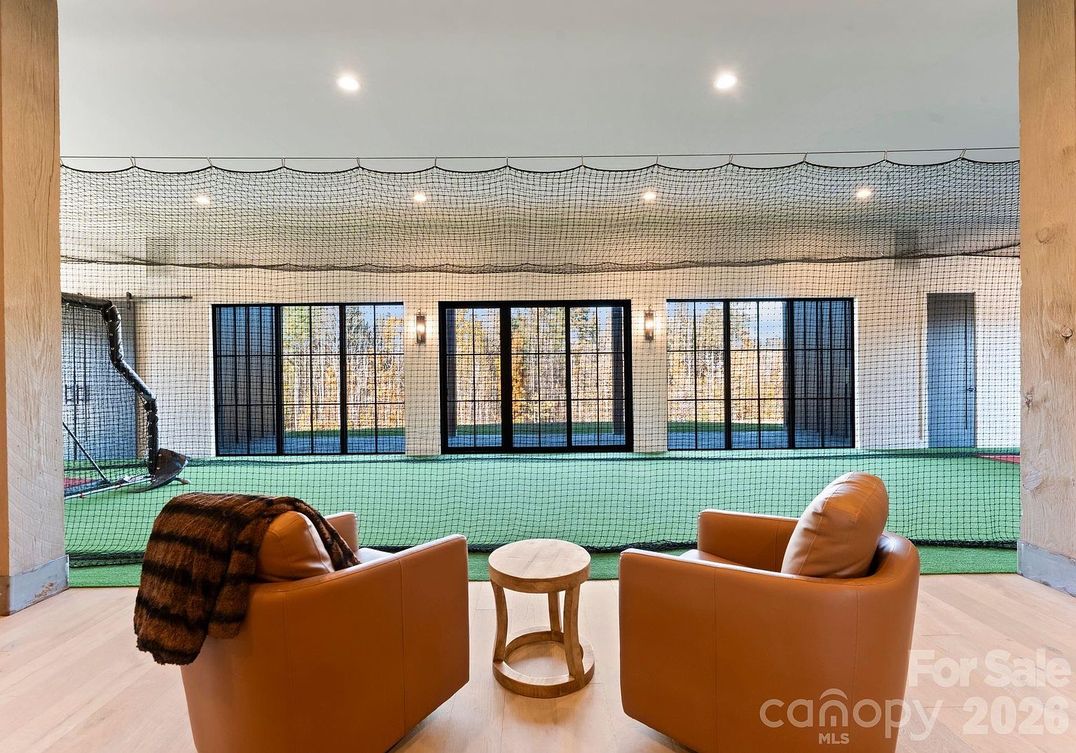 This unique interior space features a dedicated indoor sports training area enclosed by protective netting, complete with artificial turf and a batting cage setup. In the foreground, two comfortable leather armchairs and a small round side table create a relaxed viewing area, while large glass doors provide a clear view of the outdoors. The room is finished with light wood flooring and recessed lighting, offering a versatile space for athletic practice and leisure.