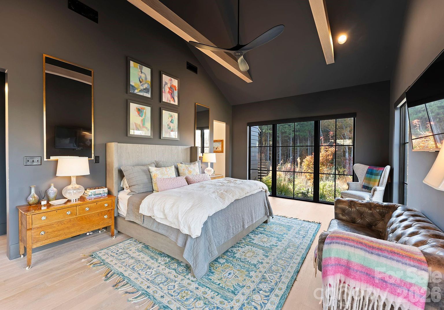 This spacious primary bedroom features a vaulted ceiling with exposed beams and a dark, moody wall color that contrasts with the light wood flooring. A large bed with a neutral upholstered headboard serves as the focal point, complemented by a patterned area rug and a cozy seating area with a tufted leather sofa. Large windows provide an expansive view of the wooded exterior, filling the room with natural light.