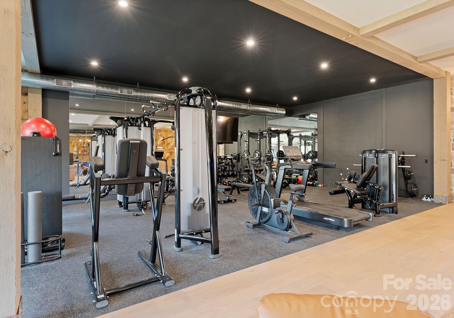 This professional-grade home gym features a dark, moody aesthetic with black ceilings, recessed lighting, and mirrored walls that enhance the sense of space. The room is equipped with a variety of fitness equipment, including a cable machine, a stationary bike, a treadmill, and a weight bench, all set upon a durable grey gym floor. The space is framed by light wood structural elements, creating a sophisticated contrast between the industrial gym equipment and the warm, natural wood tones.