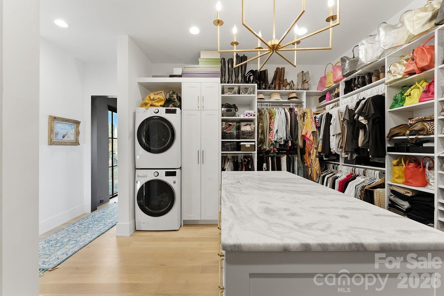 This spacious walk-in closet features a central island with a stone countertop and a built-in laundry station with stacked appliances. The room is organized with extensive shelving, hanging racks, and storage for shoes and accessories, all illuminated by a modern gold-toned chandelier. A patterned runner leads toward a doorway, highlighting the functional and luxurious design of this dressing area.