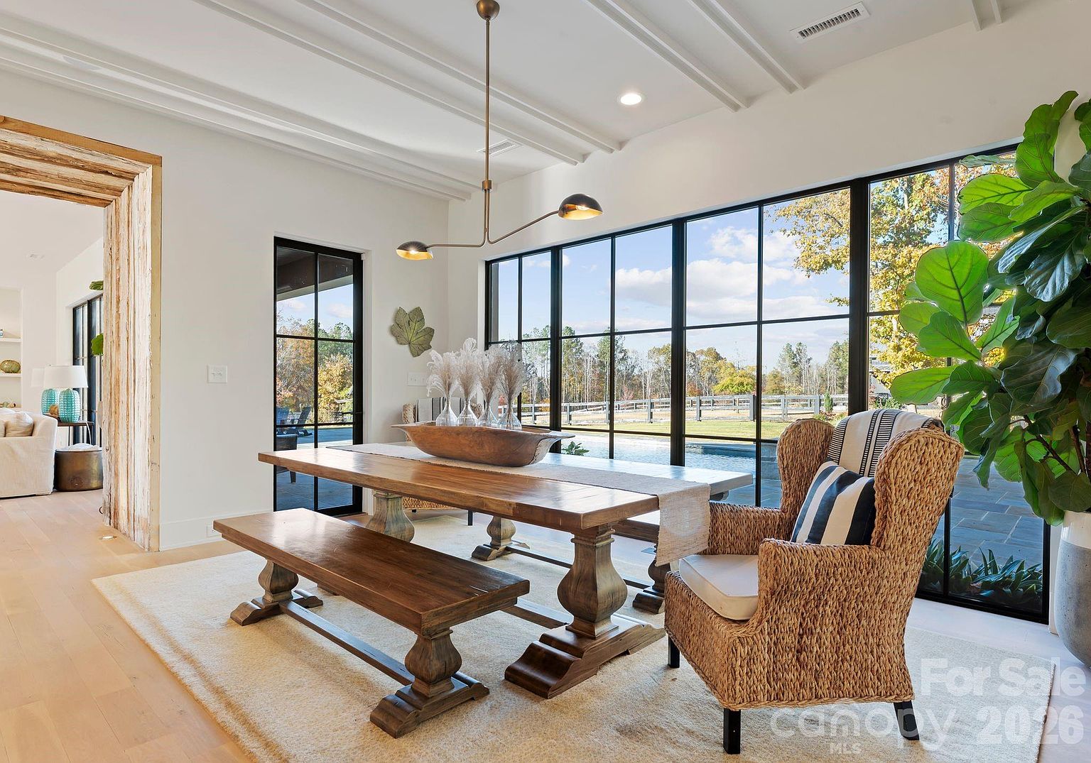 This bright and airy dining room features a large wooden table paired with a matching bench and a woven wicker armchair. The space is defined by expansive black-framed windows that offer a clear view of the outdoor pool area, creating a seamless connection between indoor and outdoor living. A modern, minimalist light fixture hangs above the table, while the neutral color palette and light wood flooring contribute to a sophisticated, transitional aesthetic.