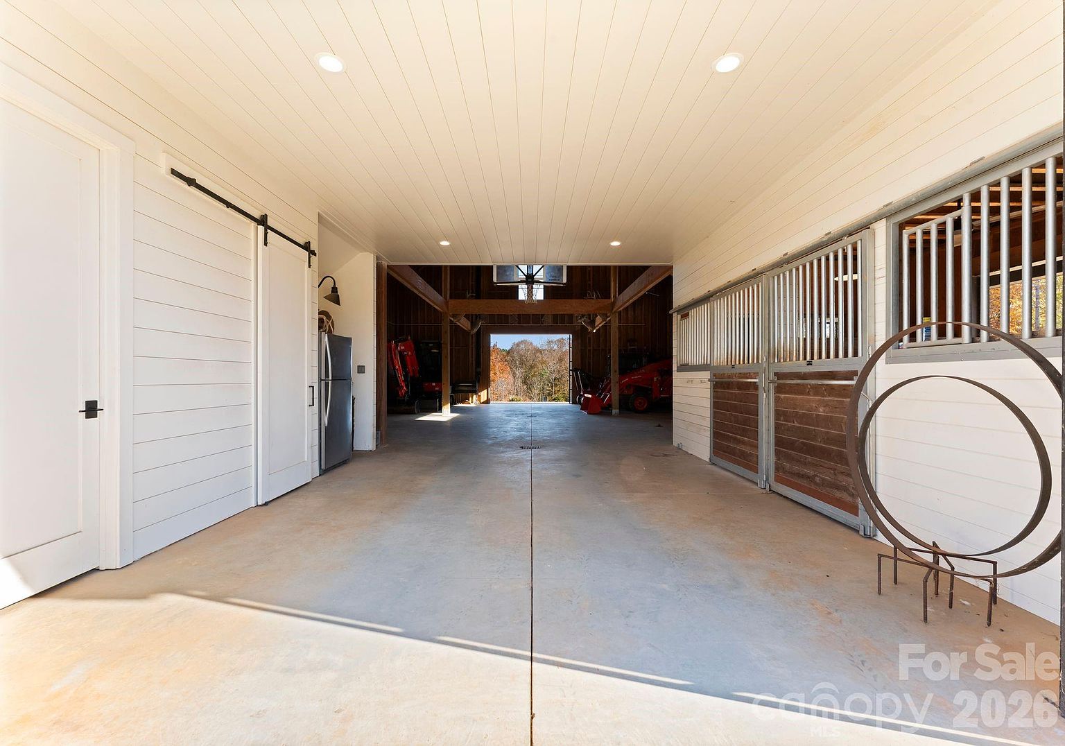 This image captures the interior of a spacious, well-lit barn or stable facility featuring a concrete floor and white shiplap walls. To the right, there are several horse stalls with metal grilles and wood-paneled lower sections, while a sliding barn door and a refrigerator are positioned on the left. The perspective looks through the structure toward an open-air barn area in the background, which houses farm equipment and offers a view of the surrounding landscape.