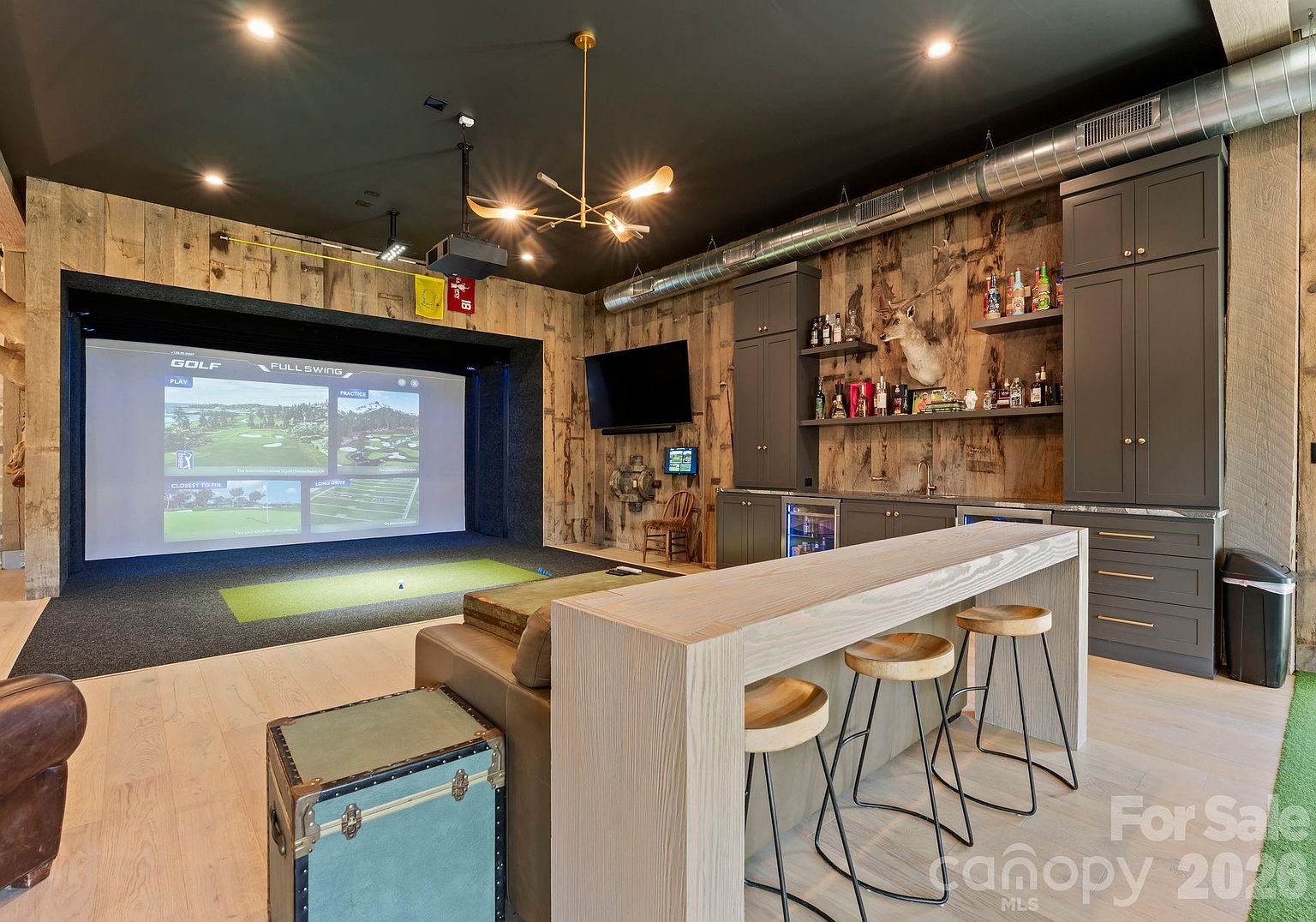 This high-end home entertainment space features a professional-grade golf simulator with a large projection screen and a dedicated hitting area. The room is designed with a rustic-industrial aesthetic, showcasing reclaimed wood wall paneling, a dark ceiling with exposed ductwork, and a built-in wet bar with dark cabinetry and open shelving. A central bar-height table with three stools provides a comfortable viewing area, creating an ideal environment for recreation and social gatherings.