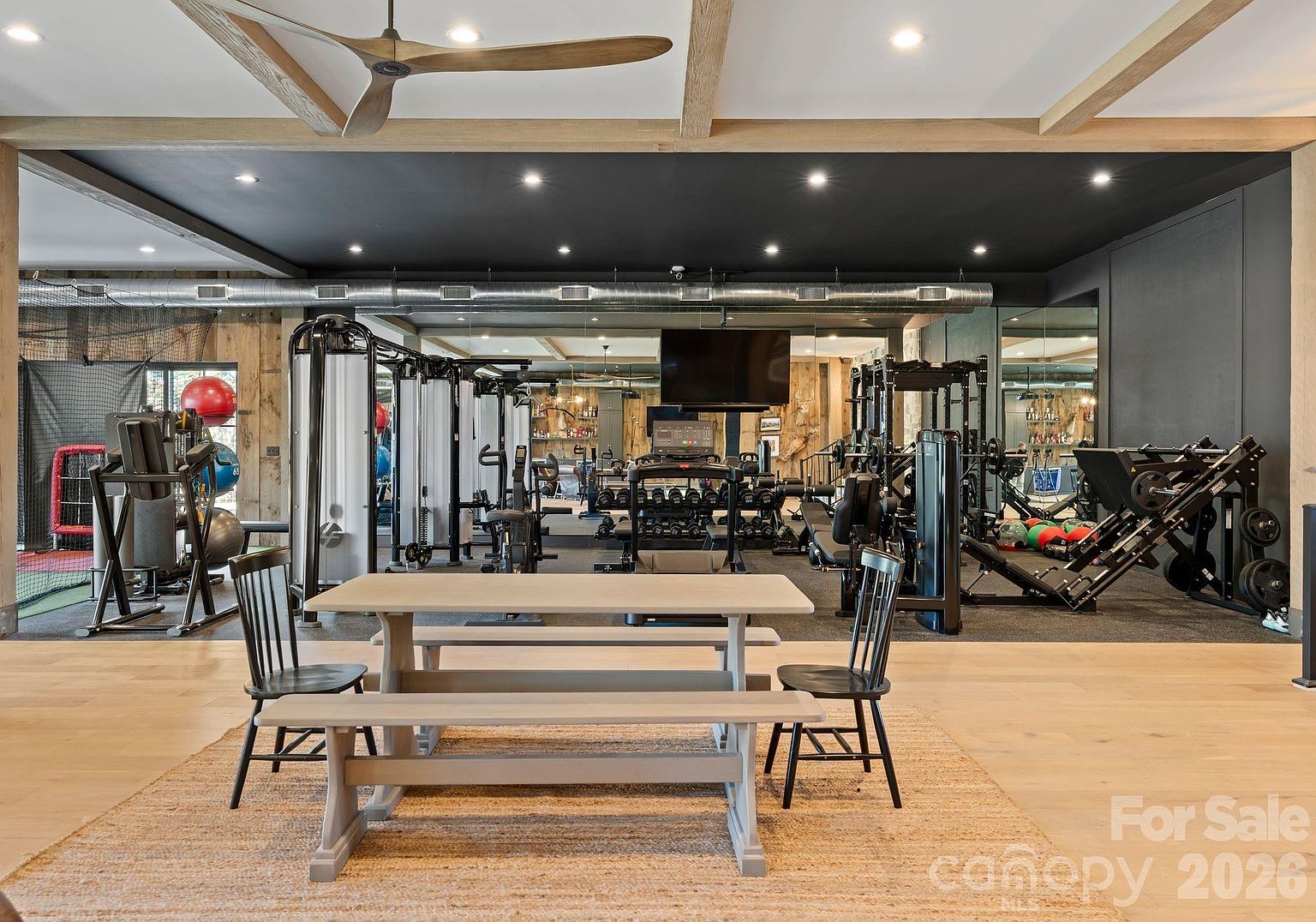 This expansive home gym features a professional-grade layout with a variety of strength training equipment, including a cable machine, a leg press, and a rack of dumbbells. The space is anchored by a central dining-style table and benches on a textured rug, creating a unique multi-functional area. The room is finished with a dark ceiling, exposed wooden beams, and mirrored walls that enhance the sense of depth and scale.