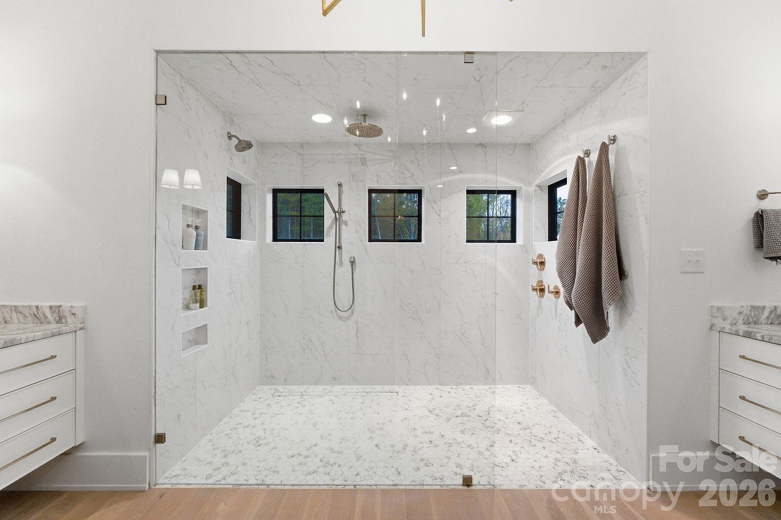 This spacious walk-in shower features light-toned stone tile walls and flooring, creating a bright and clean aesthetic. The enclosure is defined by a large glass panel, showcasing multiple shower heads, built-in wall niches for storage, and three small windows that allow natural light to filter in. Flanked by white cabinetry with gold-toned hardware on either side, the space offers a luxurious and modern spa-like atmosphere.