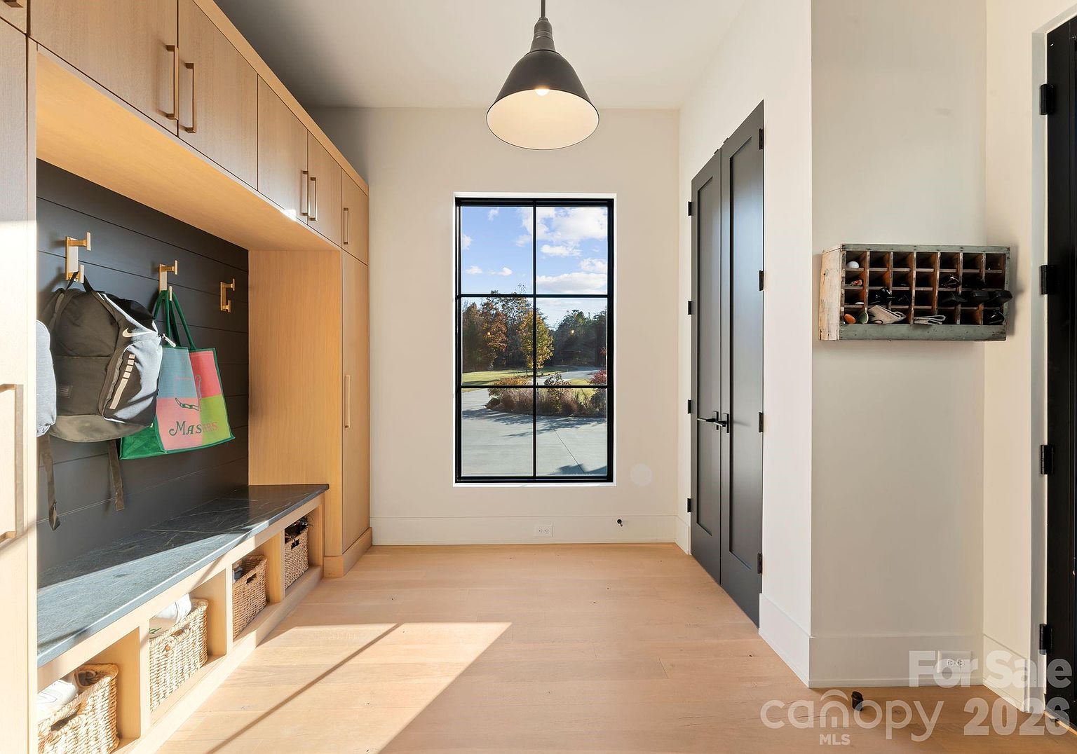 This mudroom area features custom light wood cabinetry with integrated bench seating and a dark-toned shiplap accent wall with hooks for storage. A large, black-framed window centered on the back wall provides natural light and a view of the outdoors, while a dark pendant light hangs from the ceiling. The space is finished with light-toned wood flooring and a wall-mounted vintage-style cubby organizer, creating a functional and stylish entryway.