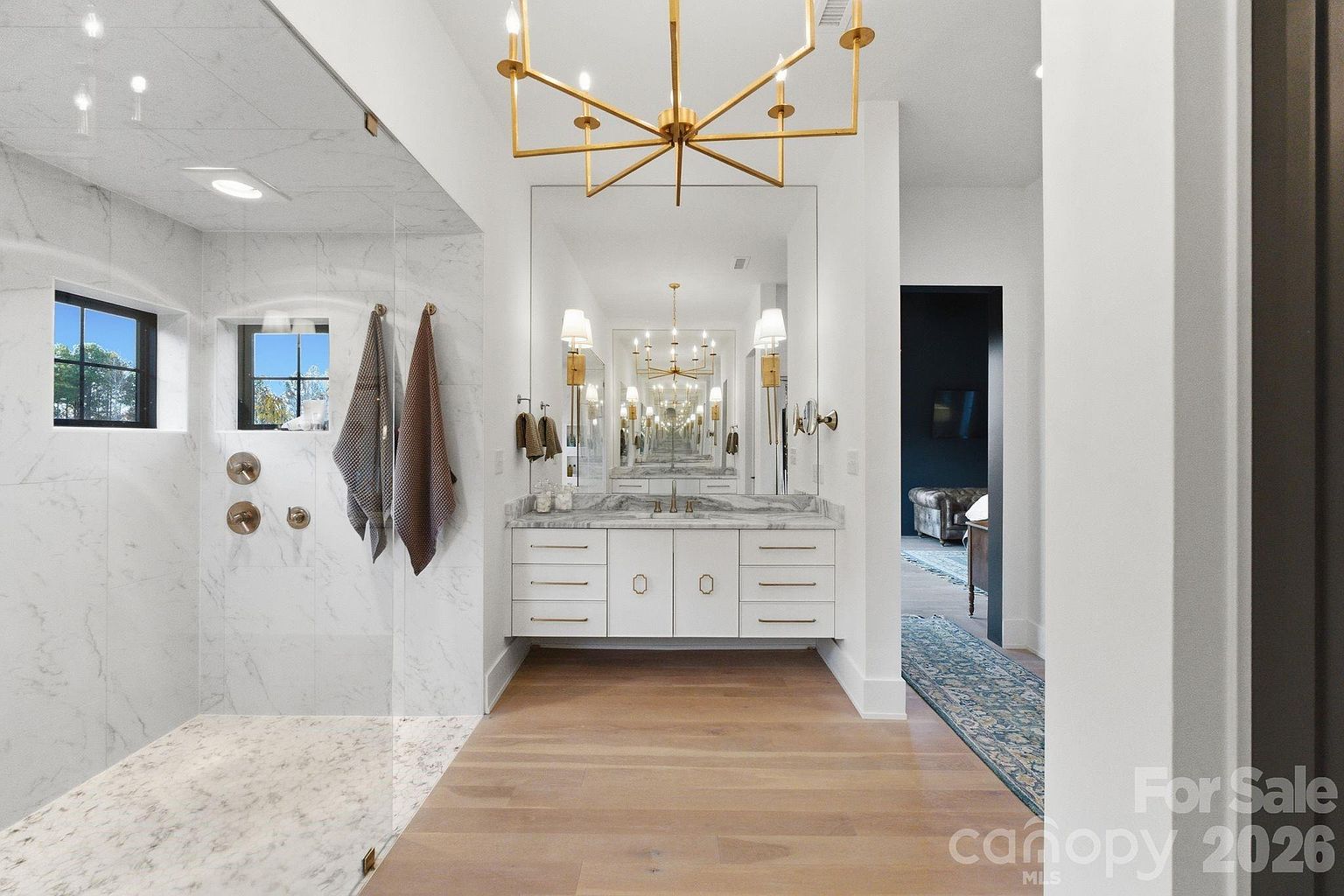 This primary bathroom features a sleek, modern design with a floating vanity topped with stone and a large, frameless glass-enclosed shower area. The space is illuminated by a prominent gold-toned chandelier and wall-mounted sconces, creating a bright and luxurious atmosphere. Light wood flooring extends throughout the room, leading toward an adjacent hallway that offers a glimpse into a dark-toned living space.