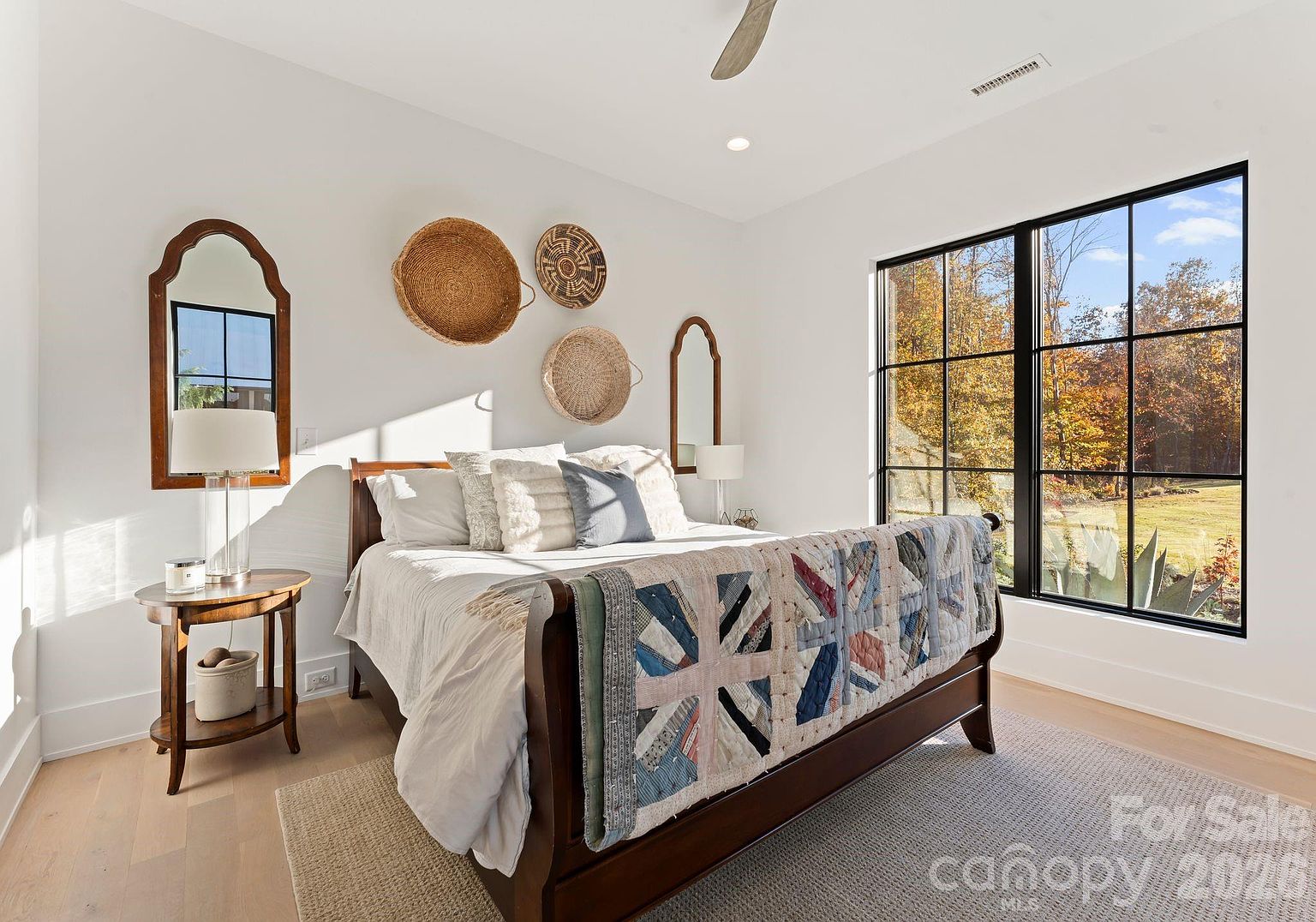 This bright and airy bedroom features a dark wood sleigh bed adorned with a colorful, patterned quilt. The room is decorated with a collection of woven wall baskets and a tall arched mirror, complemented by a round side table and a lamp. Large, black-framed windows provide an expansive view of the wooded landscape outside, filling the space with natural light.