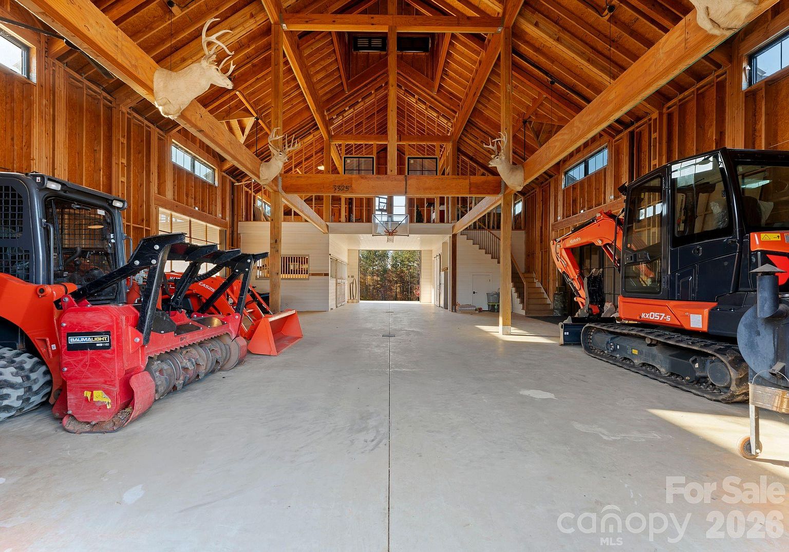 This expansive, high-ceilinged barn interior features exposed timber framing and concrete flooring, currently utilized as a large storage space for heavy machinery. The structure includes a basketball hoop mounted on a central beam, a staircase leading to an upper level, and several deer head mounts displayed on the wooden walls. The perspective is a wide, symmetrical shot looking down the center of the open floor plan toward a large, bright doorway at the far end.