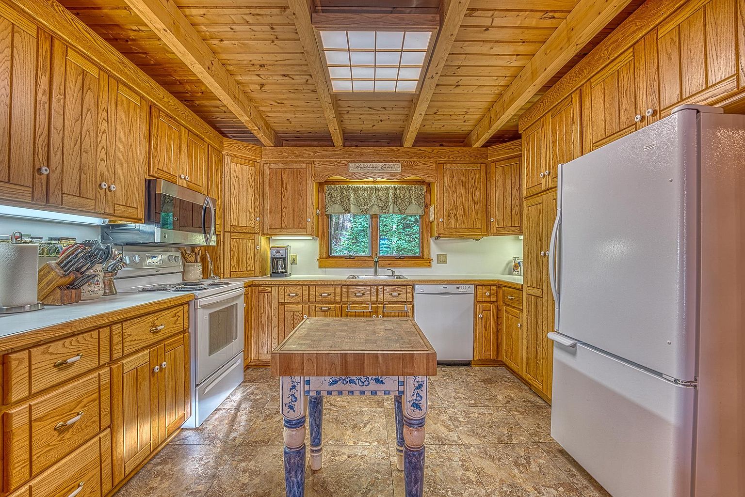 This rustic-style kitchen features warm wood cabinetry, a central butcher-block island with decorative painted legs, and a tiled floor. The space is illuminated by a large, recessed grid-style light fixture set into the wood-paneled ceiling, and a window above the sink provides a view of the outdoors. The layout includes white appliances and ample counter space, creating a cozy and functional cooking environment.