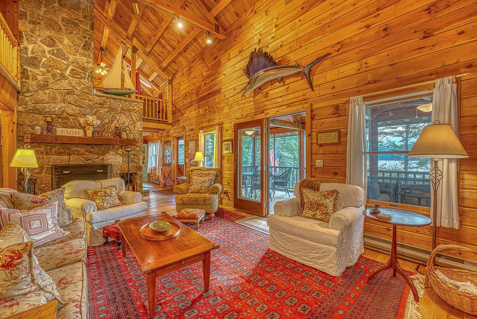 This rustic living room features warm wood-paneled walls and a vaulted ceiling, creating a cozy, cabin-like atmosphere. A prominent stone-clad chimney breast serves as a focal point, complemented by a patterned area rug and comfortable seating arranged around a wooden coffee table. Large windows and glass doors provide natural light and a view of the outdoor deck, while a decorative sailfish mount adds a unique nautical touch to the space.