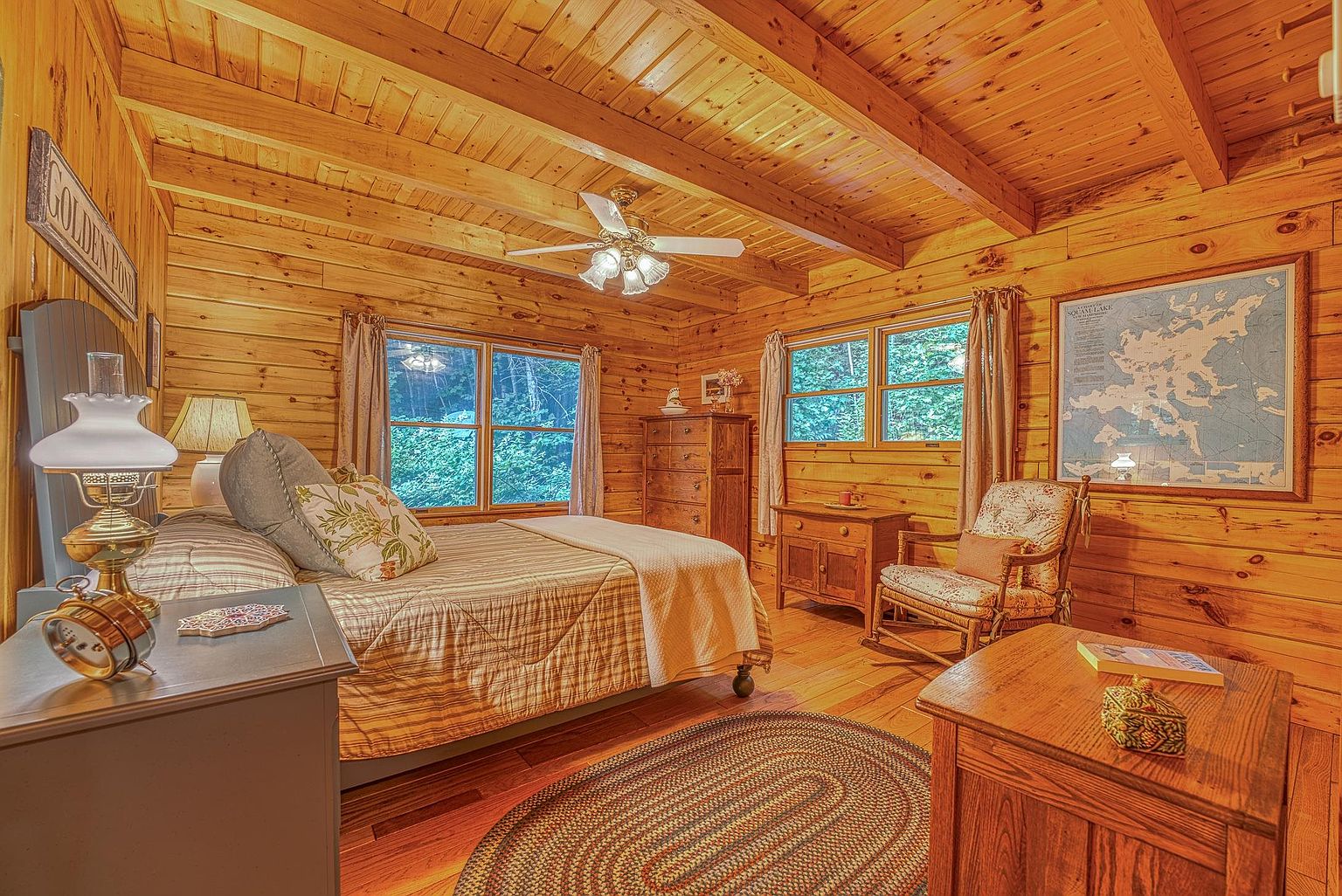 This cozy bedroom features warm, natural wood-paneled walls and a beamed ceiling, creating a rustic cabin atmosphere. The room is furnished with a comfortable bed, a wooden dresser, and a classic rocking chair, all set atop a circular braided rug. Large windows allow natural light to filter in, highlighting the wood textures and the framed map on the wall.