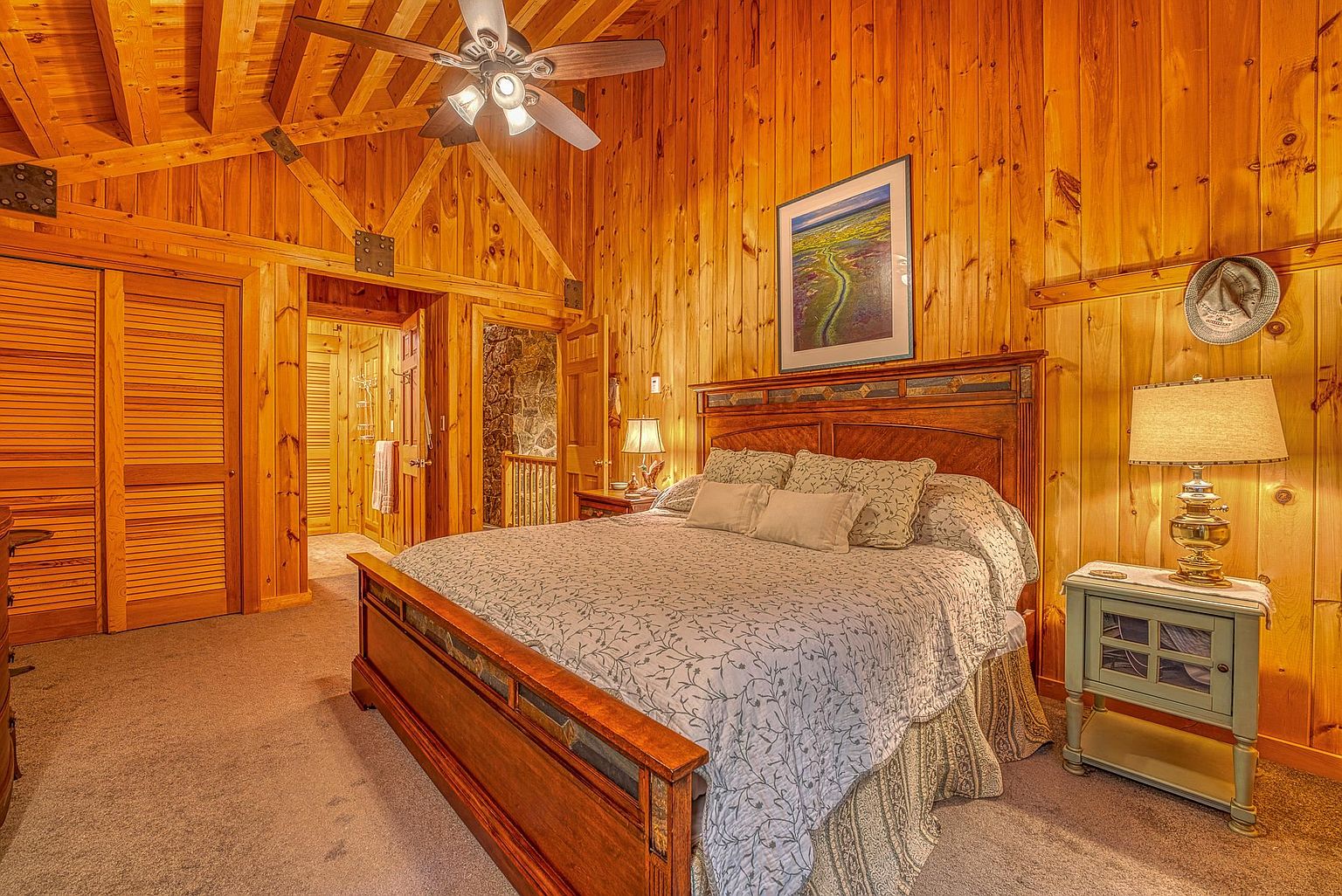 This rustic primary bedroom features warm wood-paneled walls and a vaulted ceiling with exposed beams and a ceiling fan. A large wooden bed frame with a patterned quilt serves as the focal point, complemented by a light-colored nightstand and a framed landscape print. The room includes a carpeted floor and provides a view into an adjacent hallway and a stone-walled space, creating a cozy, cabin-like atmosphere.