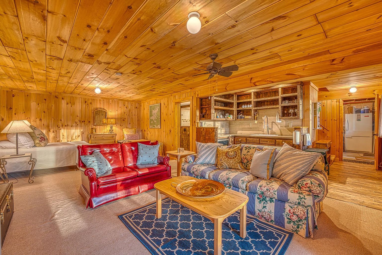This rustic-style living area features warm wood-paneled walls and ceilings, creating a cozy, cabin-like atmosphere. The space is furnished with a vibrant red leather sofa and a floral-patterned sofa, centered around a wooden coffee table on a blue patterned rug. In the background, a kitchenette with open shelving and a sink is visible, alongside a glimpse of a bedroom area, showcasing an open-concept layout.
