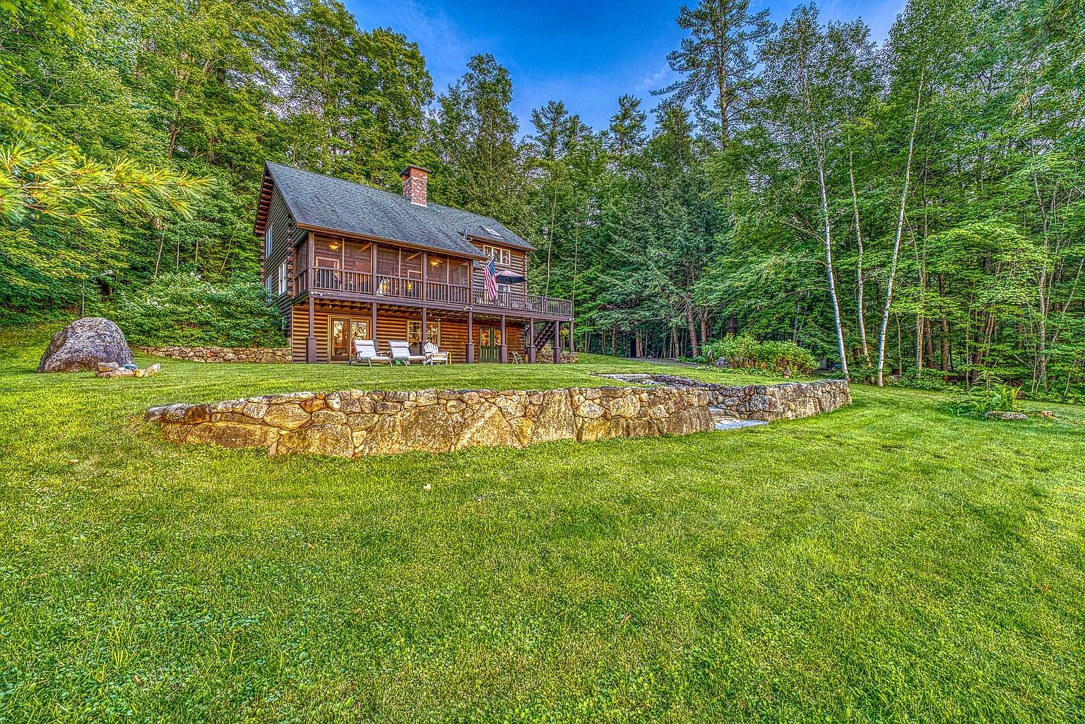 This image captures the rear exterior of a rustic log cabin nestled against a dense, lush forest backdrop. The home features a prominent wrap-around deck with a screened-in section, a stone-clad chimney, and a large, manicured lawn defined by a decorative stone retaining wall. The perspective is from a low angle on the lawn, emphasizing the peaceful, secluded nature of the property.