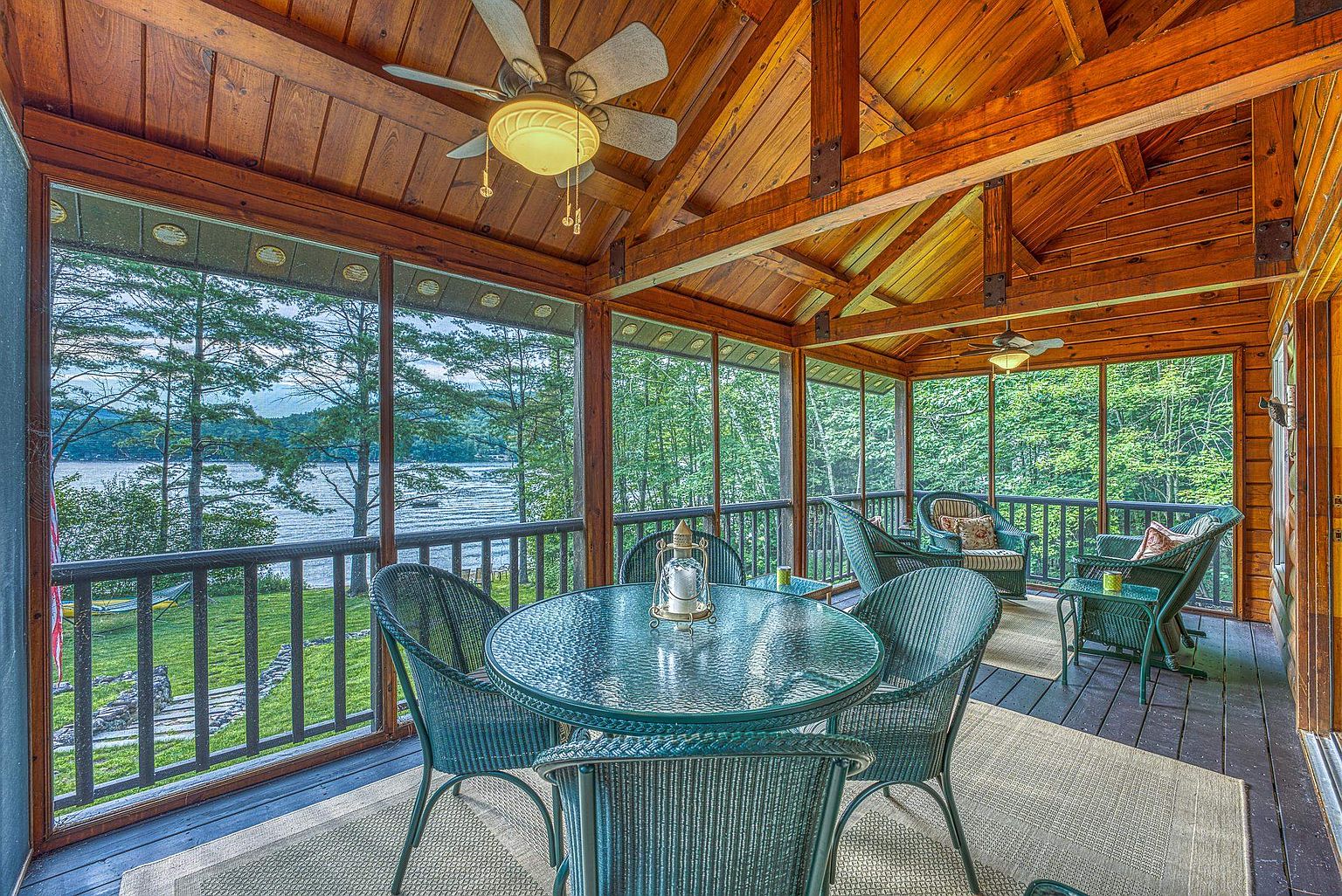 This inviting screened-in porch features a rustic, wood-paneled ceiling with exposed beams and a ceiling fan, creating a warm and cozy atmosphere. The space is furnished with a glass-topped dining set and comfortable wicker seating, all arranged to take advantage of the serene lake views visible through the large screen panels. The natural wood walls and dark decking contribute to a classic cabin aesthetic, perfect for relaxing or outdoor dining.