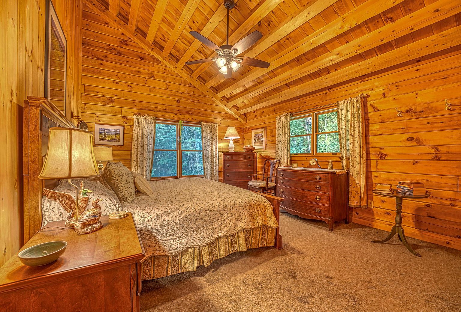 This cozy bedroom features warm wood-paneled walls and a vaulted ceiling, creating a rustic cabin atmosphere. The room is furnished with a large bed, a wooden dresser, and a small side table, all complemented by soft, neutral-toned bedding and patterned curtains. Large windows allow natural light to filter in, providing a serene view of the surrounding trees.