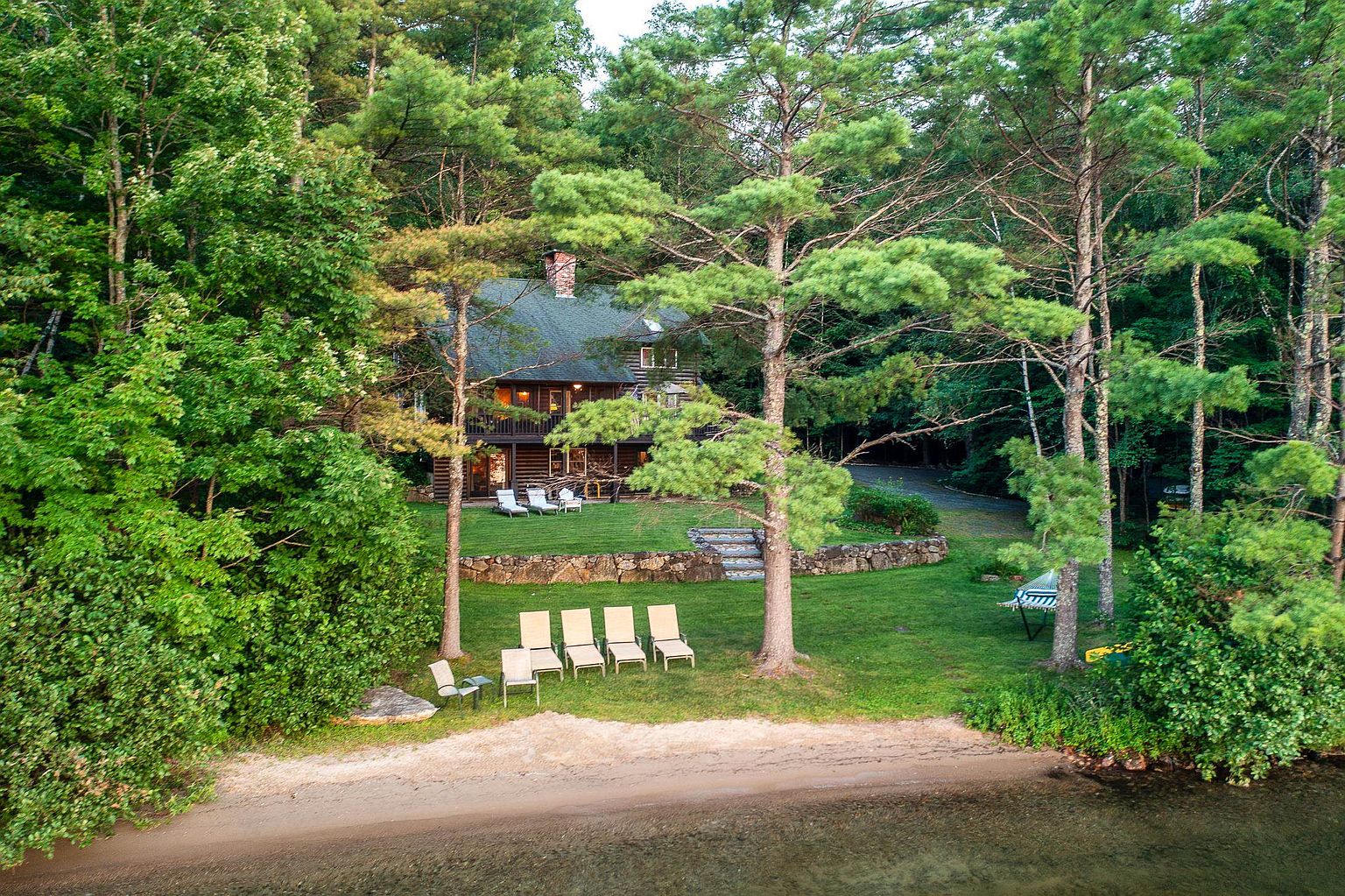 This aerial perspective captures a rustic log cabin nestled within a dense, lush forest, directly overlooking a serene lakefront. The property features a tiered lawn with a stone retaining wall, multiple outdoor seating areas, and a sandy beach shoreline. The scene conveys a peaceful, secluded retreat atmosphere, highlighting the seamless integration of the home into its natural woodland surroundings.