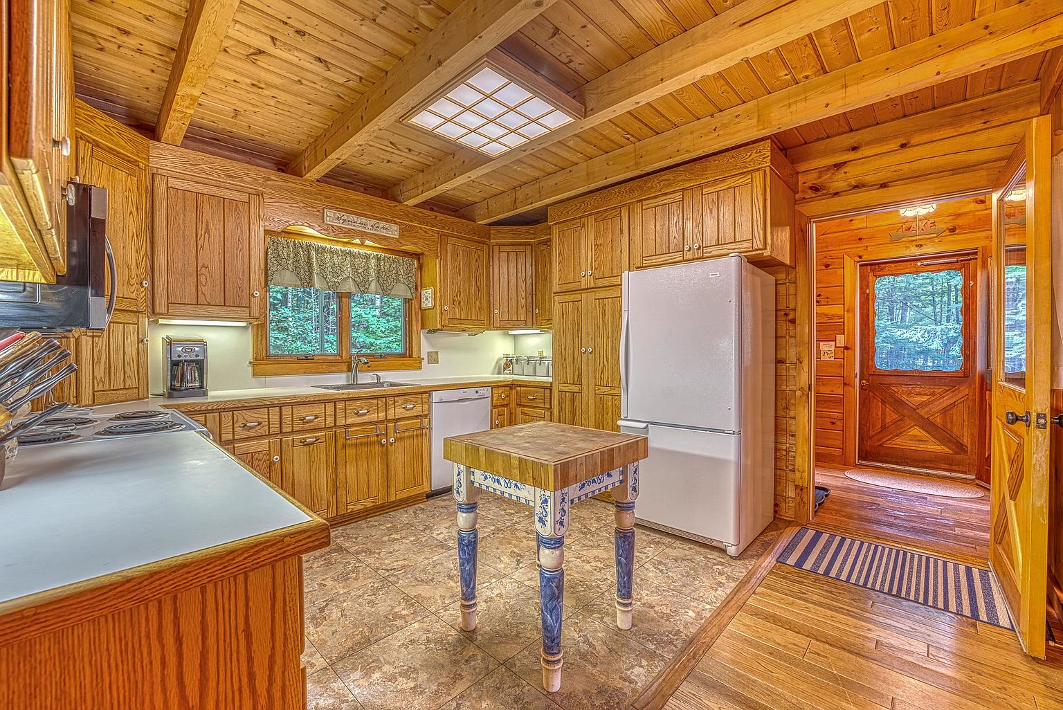 This rustic kitchen features warm wood cabinetry and exposed wooden ceiling beams, creating a cozy, cabin-like atmosphere. A central butcher-block island with decorative blue-painted legs serves as a focal point, complemented by light-toned countertops and a tiled floor. The space is well-lit by a recessed skylight and includes a white refrigerator, a dishwasher, and a window overlooking the surrounding greenery.
