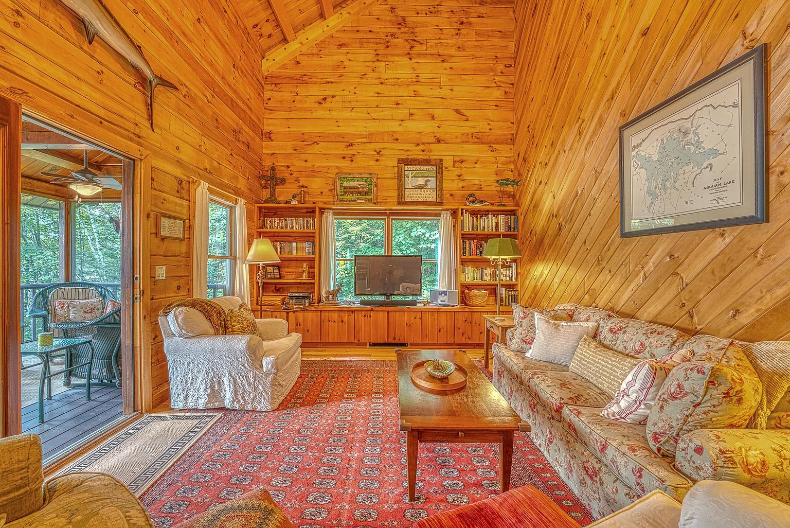 This cozy, rustic living room features warm wood-paneled walls and a vaulted ceiling, creating a cabin-like atmosphere. The space is furnished with a floral-patterned sofa, a comfortable armchair, and a central wooden coffee table set atop a large, patterned area rug. A built-in shelving unit surrounds a television, while a sliding glass door provides a view into an adjacent screened-in porch, enhancing the home's connection to the outdoors.