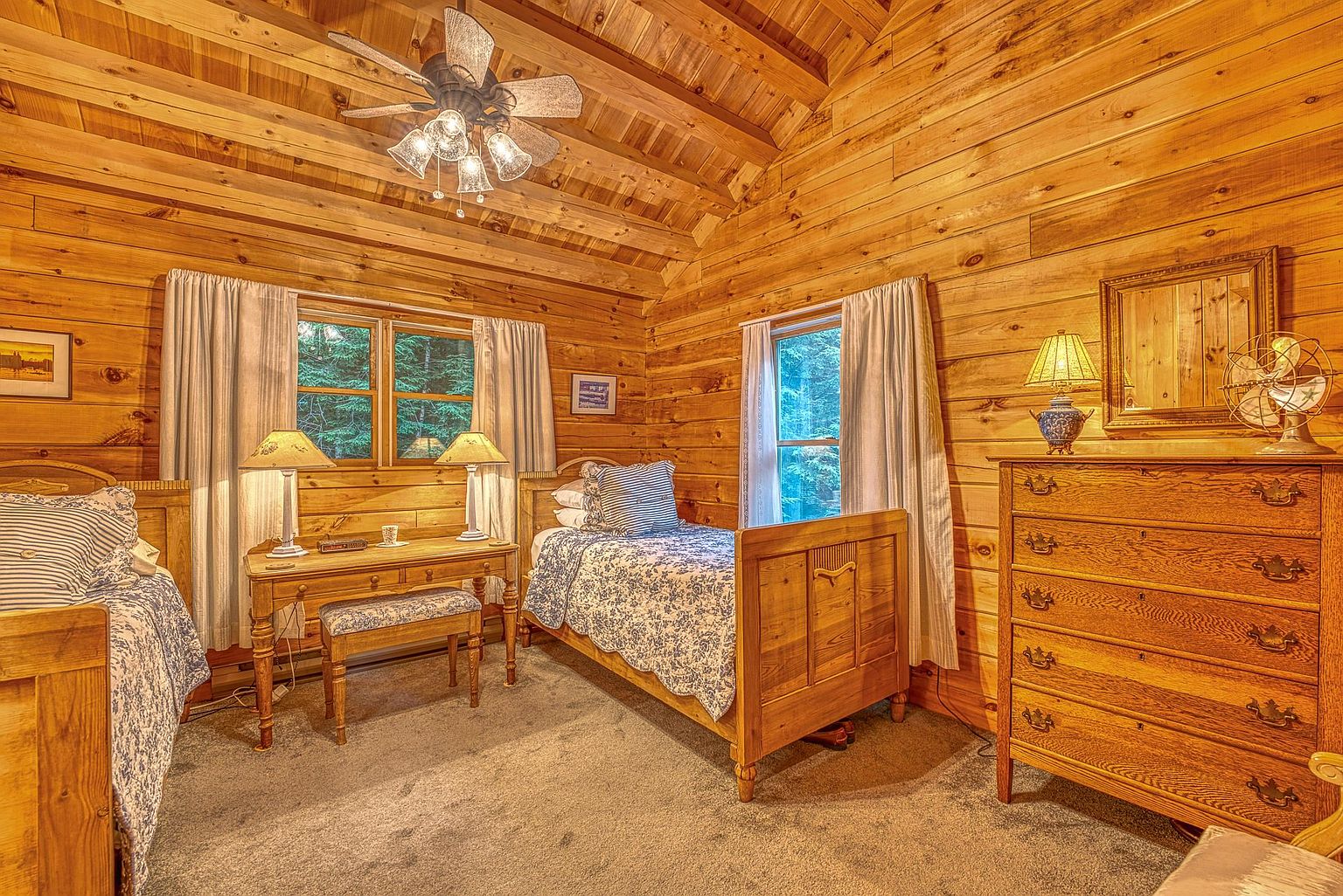 This cozy guest bedroom features warm wood-paneled walls and a vaulted ceiling, creating a rustic cabin atmosphere. The room is furnished with two twin beds, a central wooden desk with a matching bench, and a large wooden dresser topped with a lamp and a decorative fan. The space is well-lit by natural light streaming through windows dressed in simple curtains, and the overall aesthetic is traditional and inviting.
