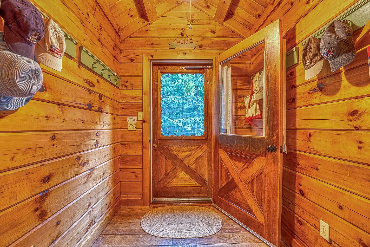 This rustic entryway features warm, horizontal wood-paneled walls and a matching wood door with a glass insert that looks out onto a lush, green forest. A circular woven rug sits on the tiled floor, while a wooden coat rack holds several hats, creating a cozy and welcoming atmosphere. The perspective is centered on the doorway, highlighting the natural wood textures and the inviting transition between the interior cabin space and the outdoors.