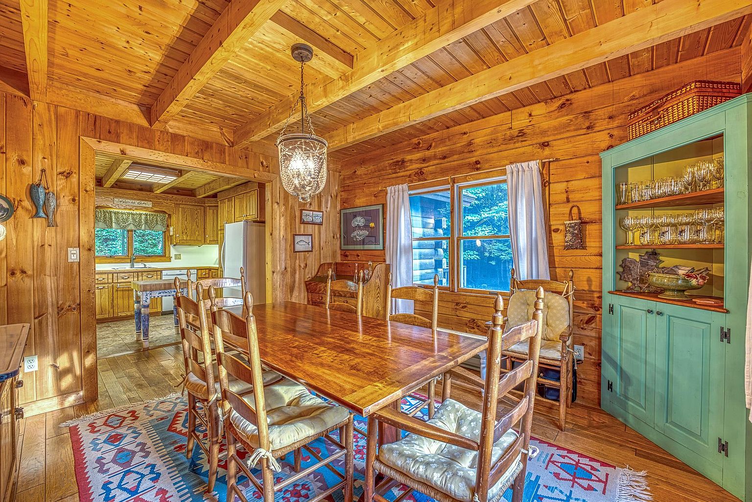 This rustic dining room features warm wood-paneled walls and exposed ceiling beams, creating a cozy, cabin-like atmosphere. A large wooden dining table with matching chairs sits atop a vibrant, patterned area rug, while a distinctive teal-painted hutch provides a pop of color and storage for glassware. The space flows seamlessly into an adjacent kitchen, with a decorative pendant light fixture hanging centrally above the table.