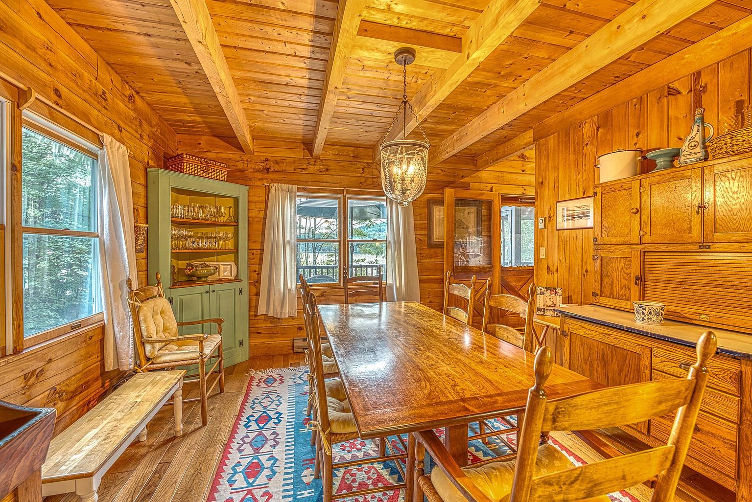 This rustic dining room features warm wood-paneled walls and exposed ceiling beams, creating a cozy, cabin-like atmosphere. A long wooden dining table serves as the centerpiece, surrounded by wooden chairs with cushioned seats, all set atop a vibrant, patterned area rug. Large windows allow natural light to filter in, while a decorative hanging pendant light adds a touch of charm to the space.