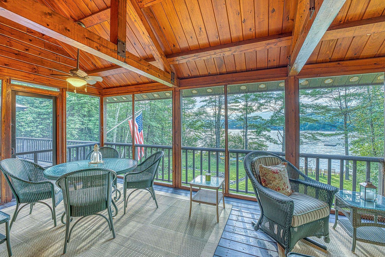 This inviting screened-in porch features a rustic aesthetic with exposed wood beams and a vaulted ceiling. The space is furnished with wicker-style seating, including a dining set and a comfortable armchair, all arranged on a neutral area rug. Large screen panels provide an unobstructed, panoramic view of the lake and surrounding trees, creating a serene and relaxing atmosphere.