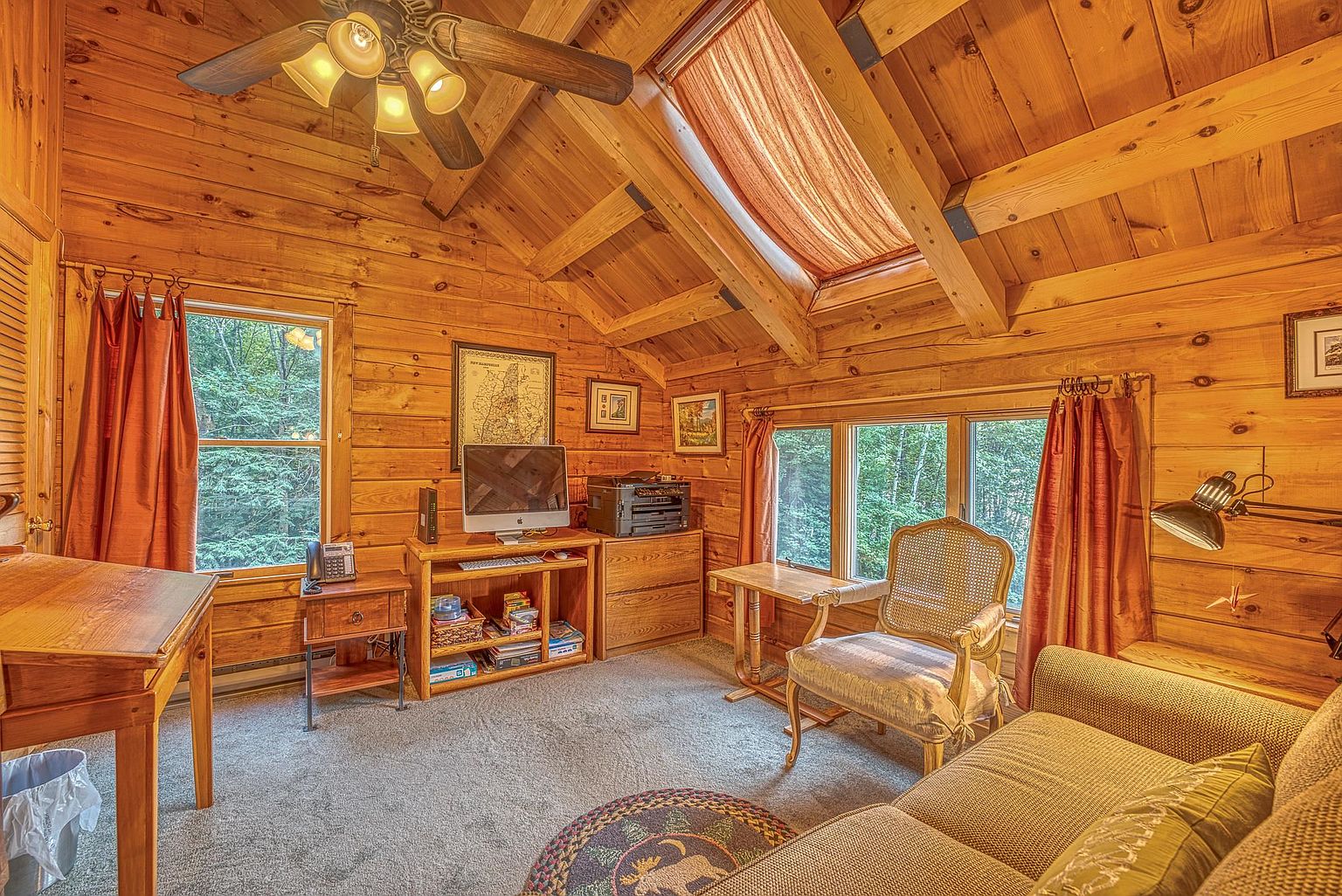 This cozy, rustic home office features warm wood-paneled walls and a vaulted ceiling with exposed beams and a skylight. The room is furnished with a computer desk, a printer station, a comfortable armchair, and a sofa, creating a functional yet inviting workspace. Large windows provide natural light and views of the surrounding trees, enhancing the cabin-like atmosphere.