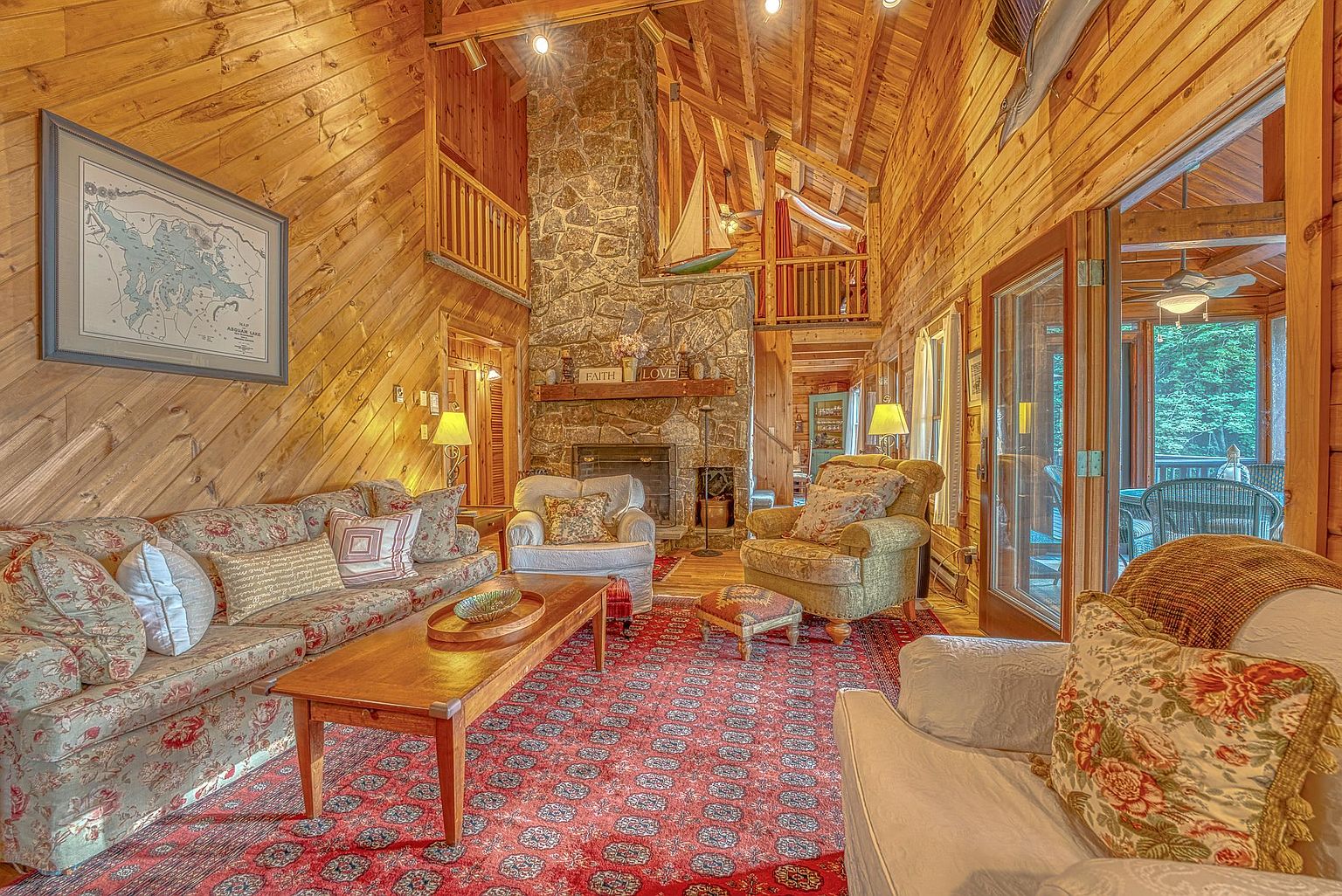 This rustic living room features warm wood-paneled walls and a vaulted ceiling with exposed beams, creating a cozy, cabin-like atmosphere. A prominent stone-clad chimney breast serves as the focal point, complemented by a large, patterned area rug and comfortable, traditional-style seating. The space is bathed in natural light from the adjacent sunroom, offering a welcoming and inviting environment.
