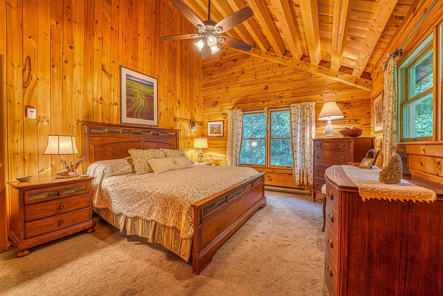 This cozy primary bedroom features warm wood-paneled walls and a vaulted ceiling with exposed beams, creating a rustic cabin aesthetic. The room is furnished with a large wooden bed frame, matching nightstands, and a dresser, all complemented by soft carpet and patterned bedding. Large windows with floral curtains allow natural light to filter in, providing a view of the surrounding trees.