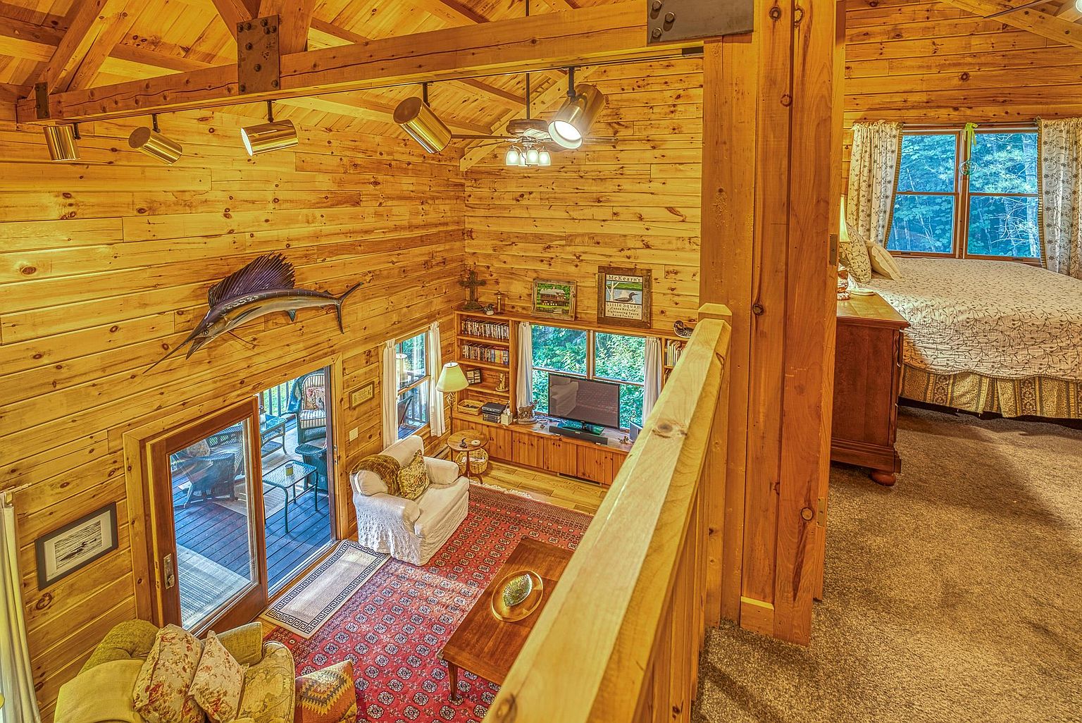 This elevated perspective captures a cozy, rustic-style living area with warm wood-paneled walls and ceilings, featuring a lofted view looking down into the main space. The room is furnished with a comfortable armchair, a patterned area rug, and a media console, while a large glass door provides access to an outdoor deck. To the right, a partially visible bedroom area with a neatly made bed adds to the inviting, cabin-like atmosphere of the home.