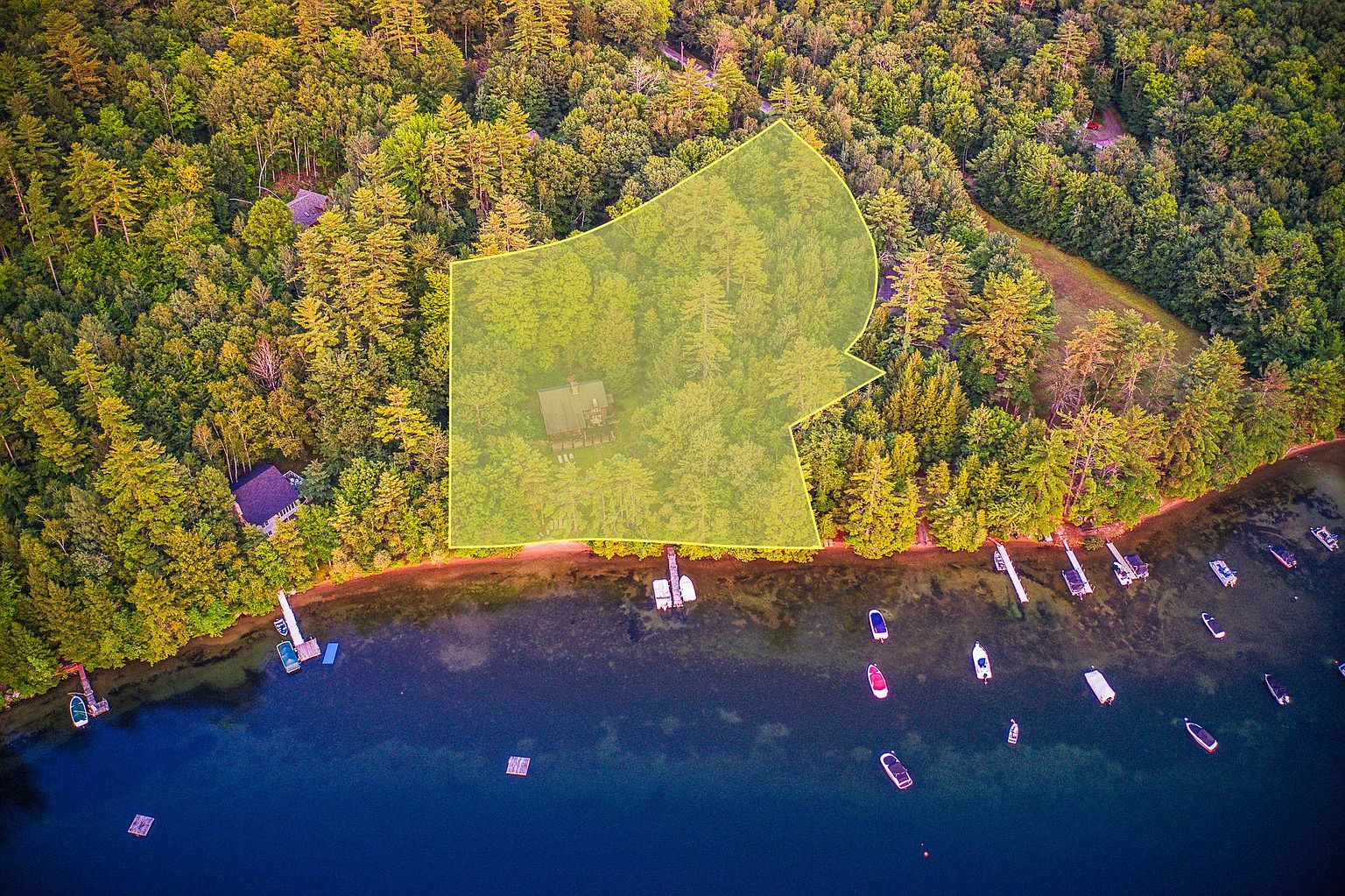 This aerial view captures a secluded waterfront property nestled within a dense, lush forest. A highlighted yellow polygon outlines the expansive wooded lot, which features a private dock extending into the calm, deep blue lake. The perspective provides a comprehensive look at the property's natural surroundings, including the shoreline, nearby boats, and the serene, wooded landscape.