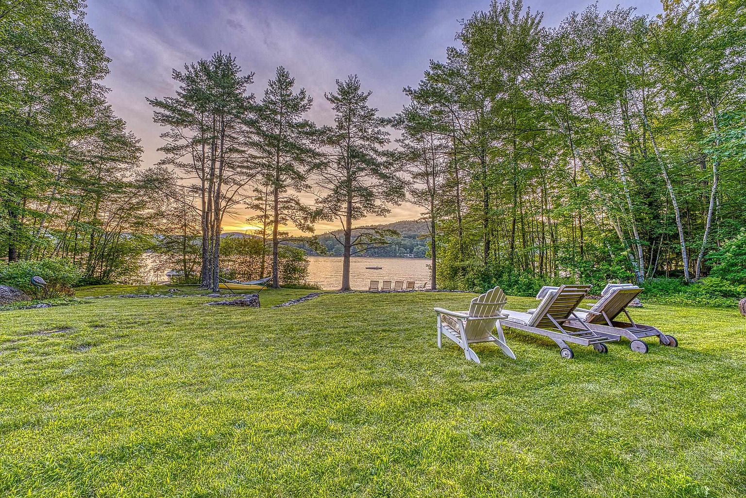 This serene backyard scene features a lush, expansive green lawn leading toward a tranquil lake framed by mature trees. In the foreground, two wooden lounge chairs are positioned to enjoy the sunset view, while a hammock is visible nestled between the trees near the water's edge. The perspective is a wide, eye-level shot that captures the peaceful, natural beauty of the property's waterfront setting.