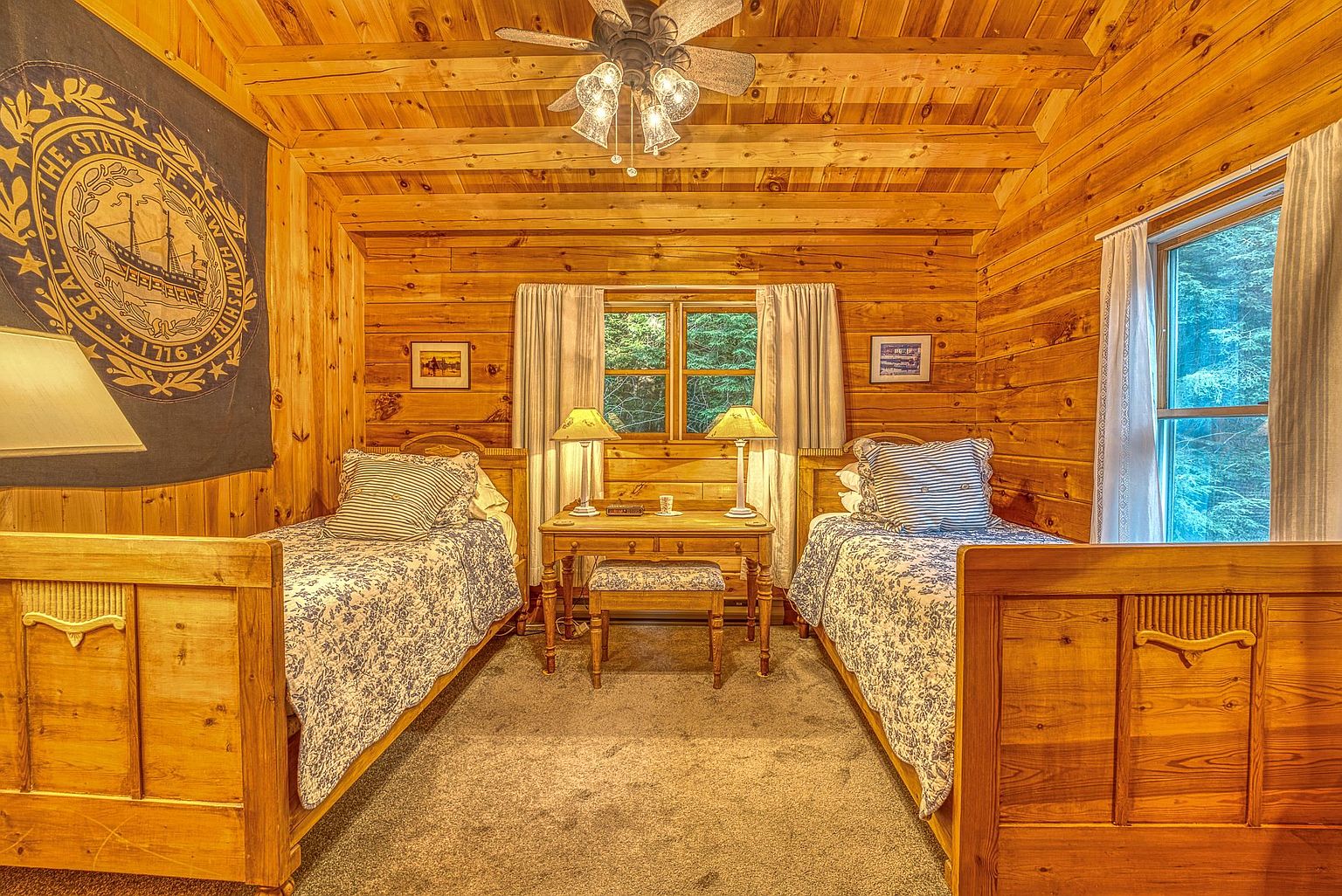 This cozy, rustic-style bedroom features two twin beds with patterned bedding and striped accent pillows, centered around a small wooden desk and stool. The room is finished with warm, horizontal wood-paneled walls and a ceiling fan, creating a cabin-like atmosphere. A large flag representing the State of New Hampshire hangs on the left wall, and windows with light curtains allow natural light to filter into the space.